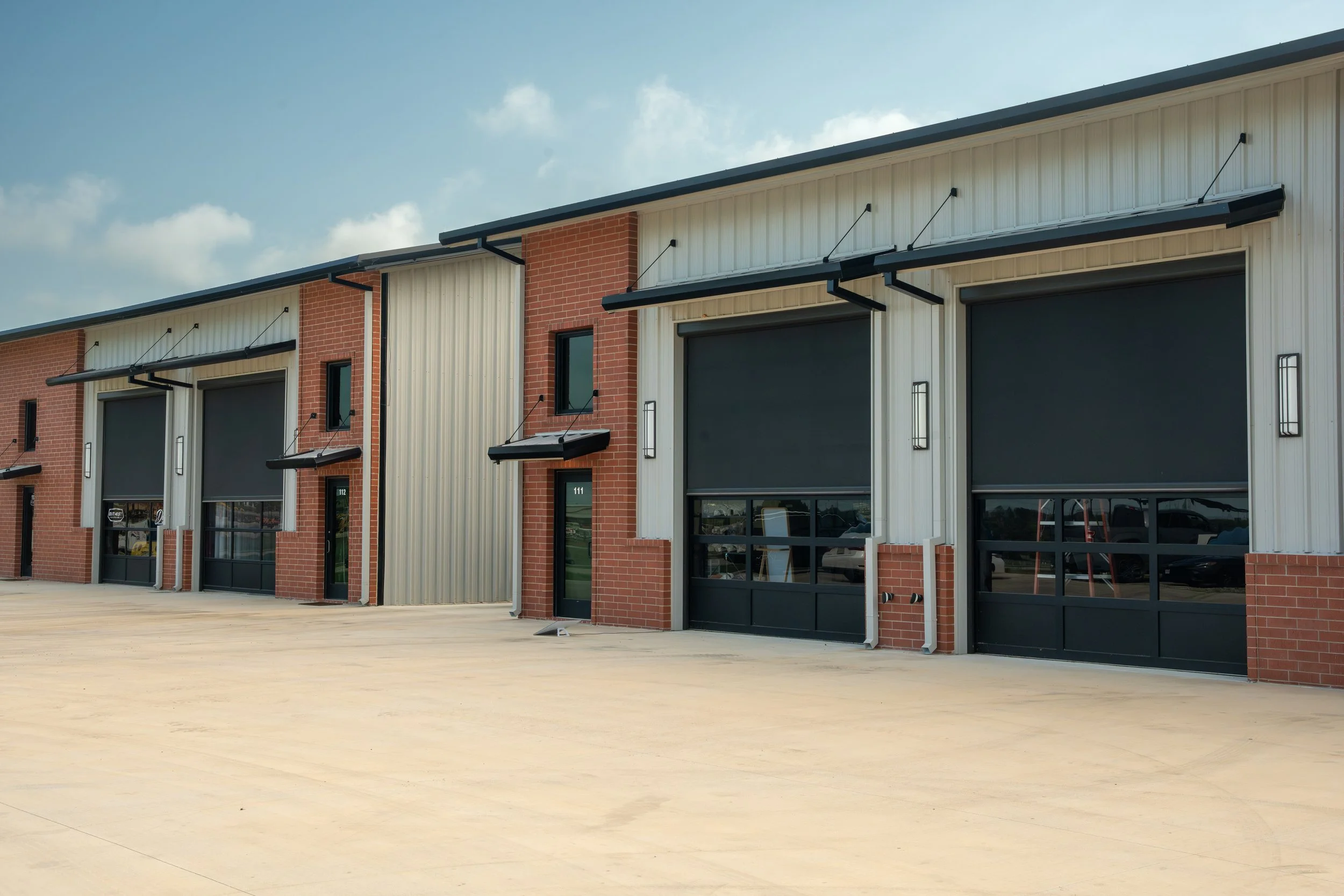 Modern commercial warehouse with black garage doors, brick and metal siding, and sidewalk in front.