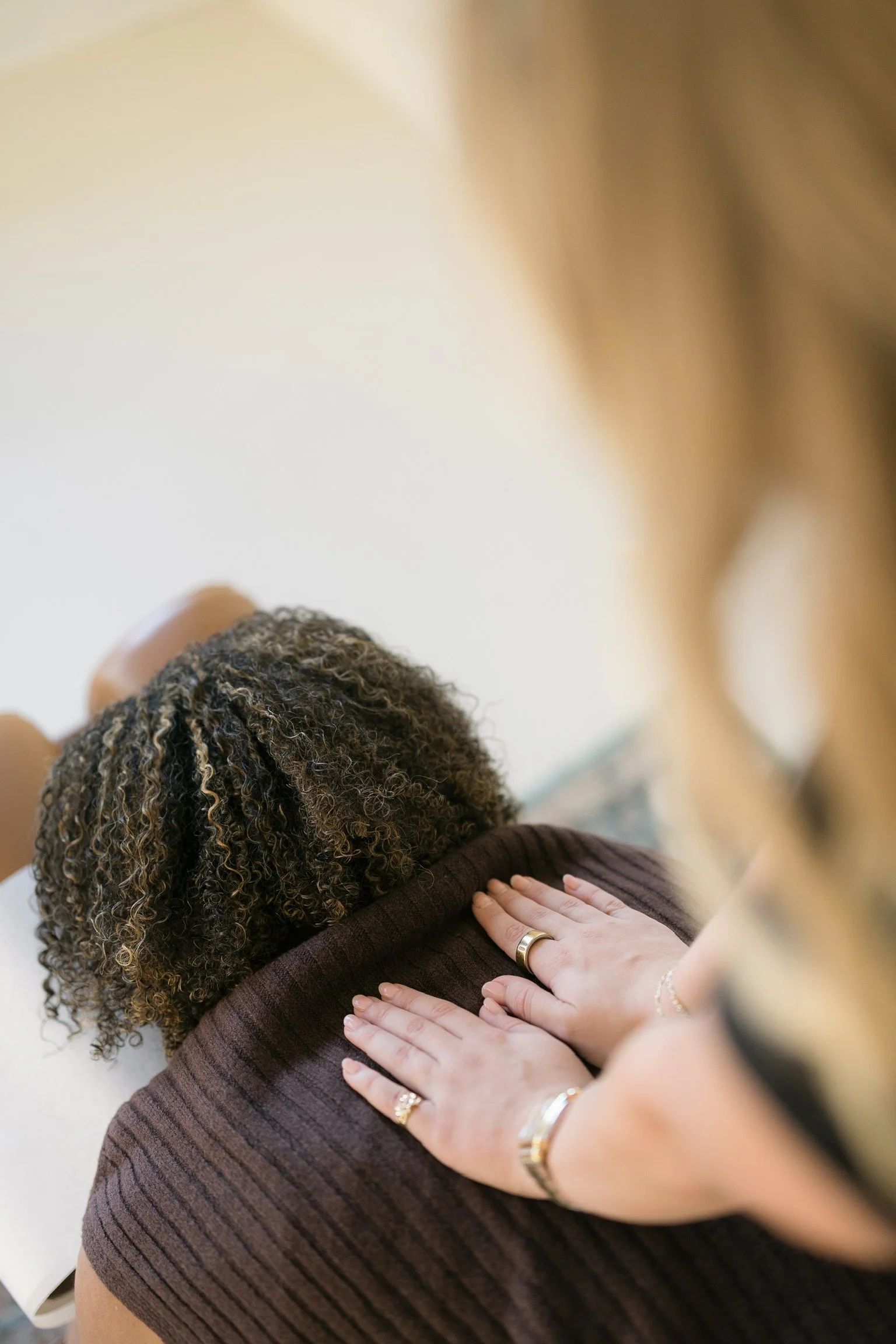 Close-up of a person's back with hands resting on their shoulder, showing rings and jewelry, with curly hair and wearing a brown ribbed shirt.