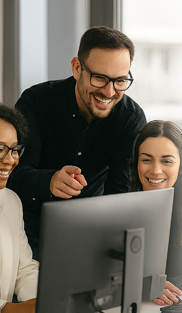 Three people gathered around a computer monitor, smiling and looking at the screen, in an office setting.