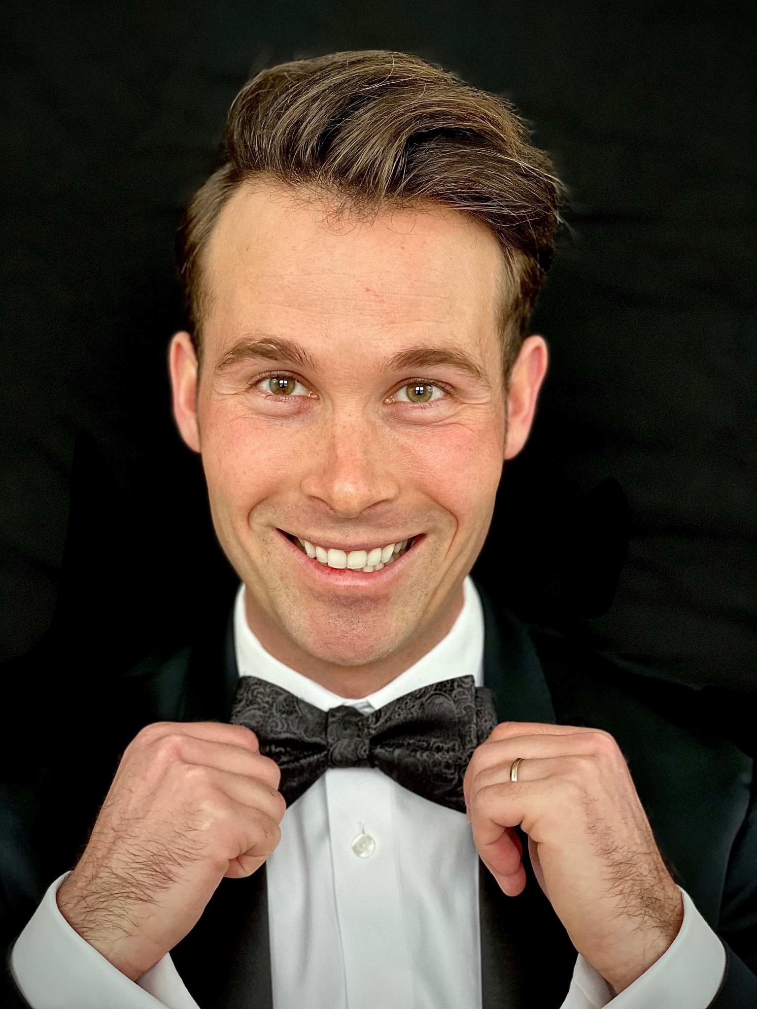 A smiling man in a tuxedo adjusting his bowtie, with a dark background.
