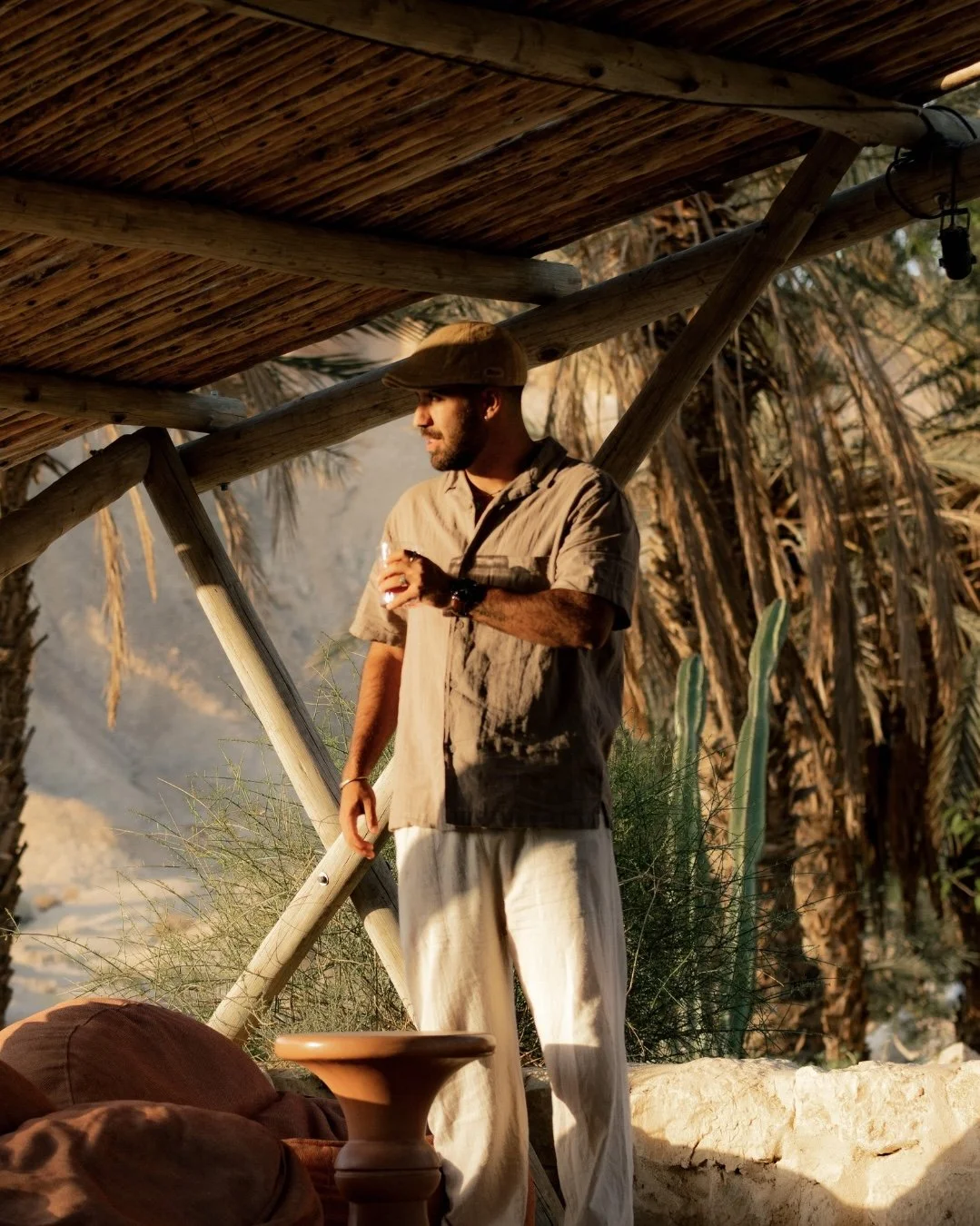 A man standing outdoors in the sunlight, holding a drink, under a makeshift wooden canopy, with desert plants and mountains in the background.