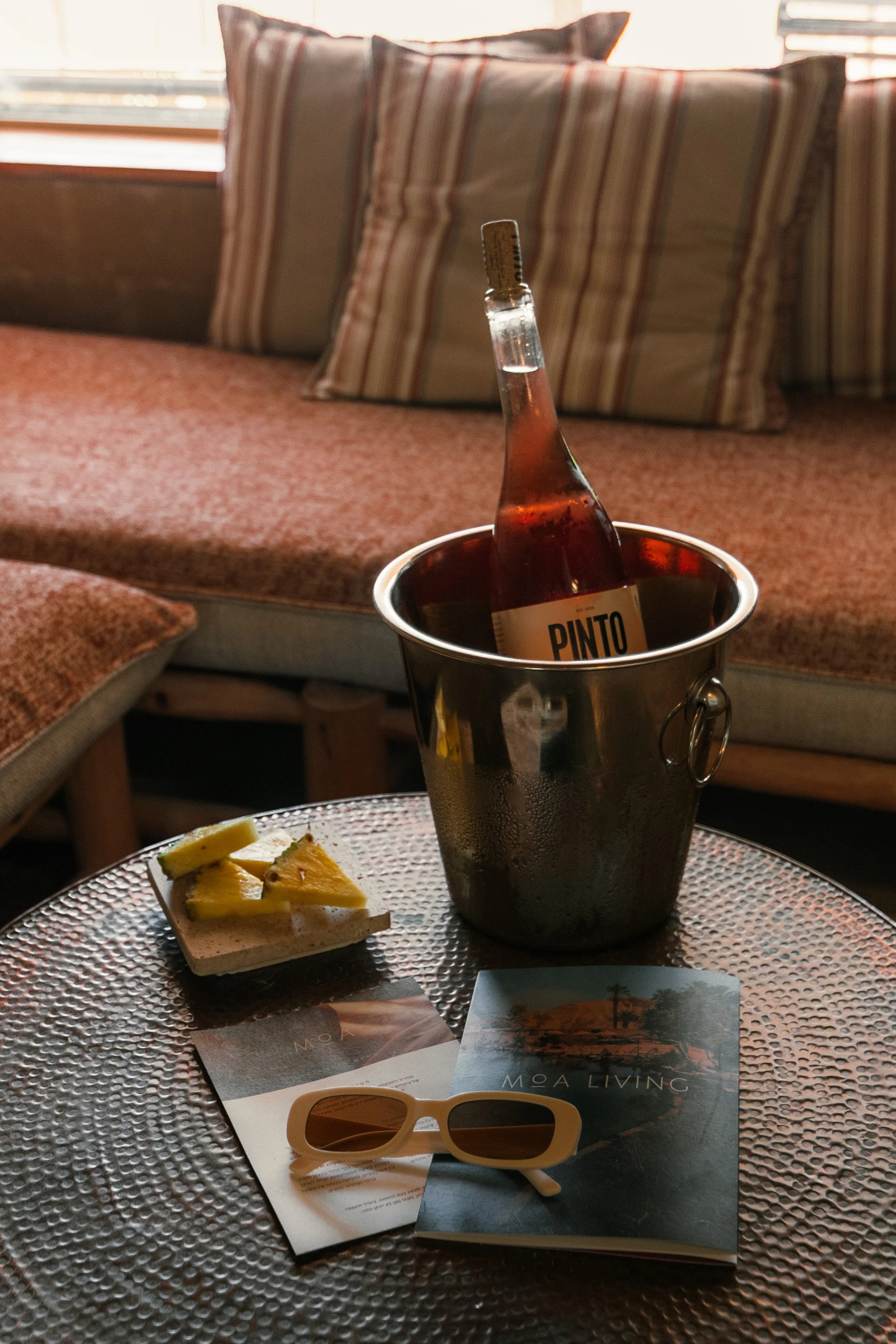 A metal ice bucket with a bottle labeled 'Pinto' inside, placed on a textured round table. The table also holds a small plate of pineapple slices, a pair of sunglasses, and a brochure or magazine. In the background, there is a cushioned bench with striped pillows.