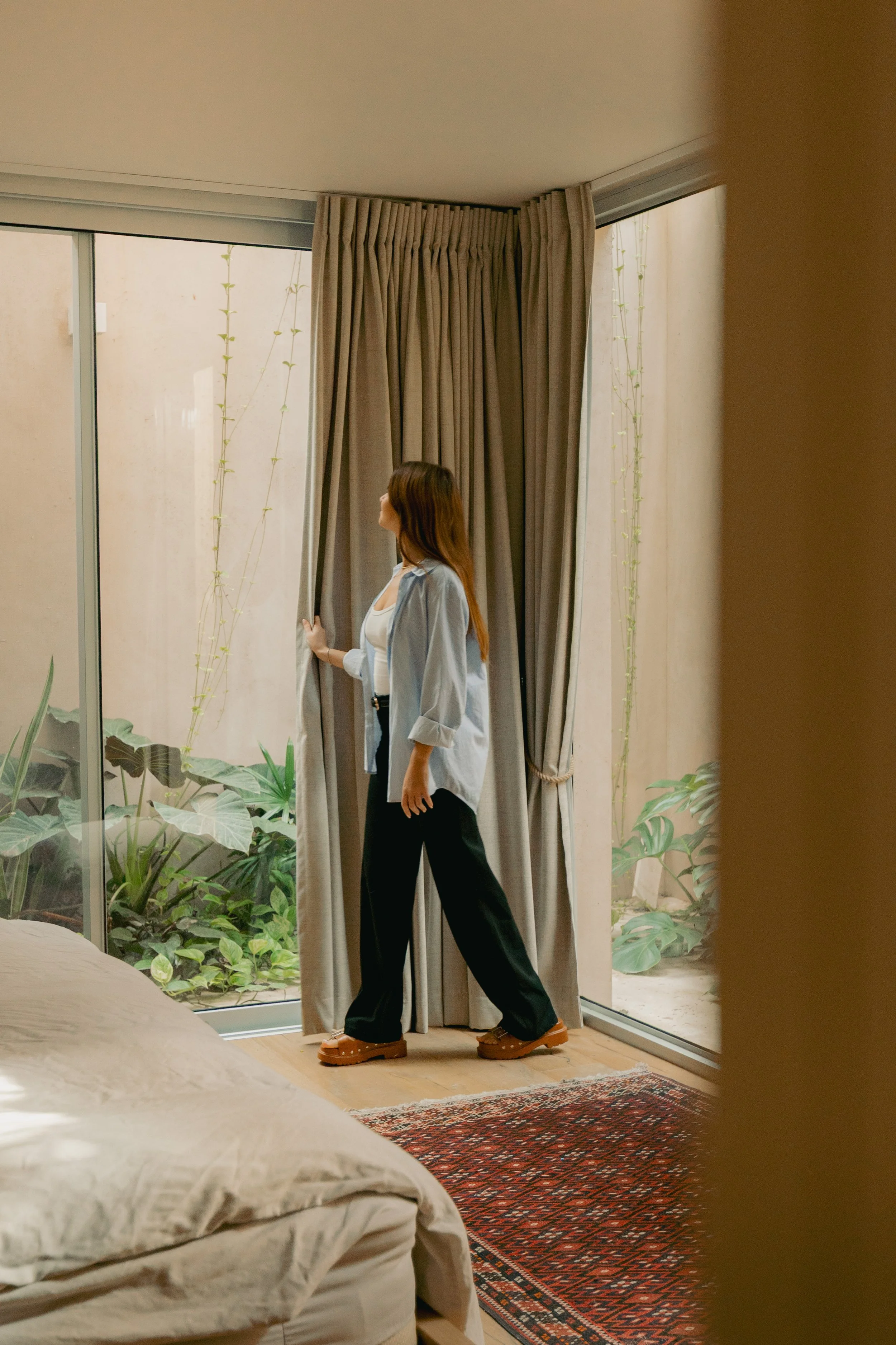 A woman standing by a large window with curtains, looking outside, with greenery visible through the glass.