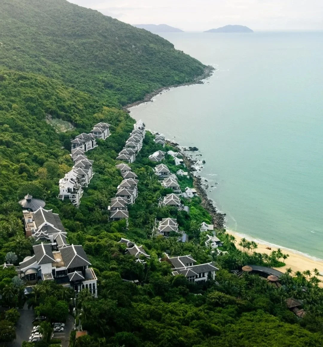 Aerial view of a coastal resort with multiple white buildings among lush greenery, overlooking a beach and ocean with small islands in the distance.