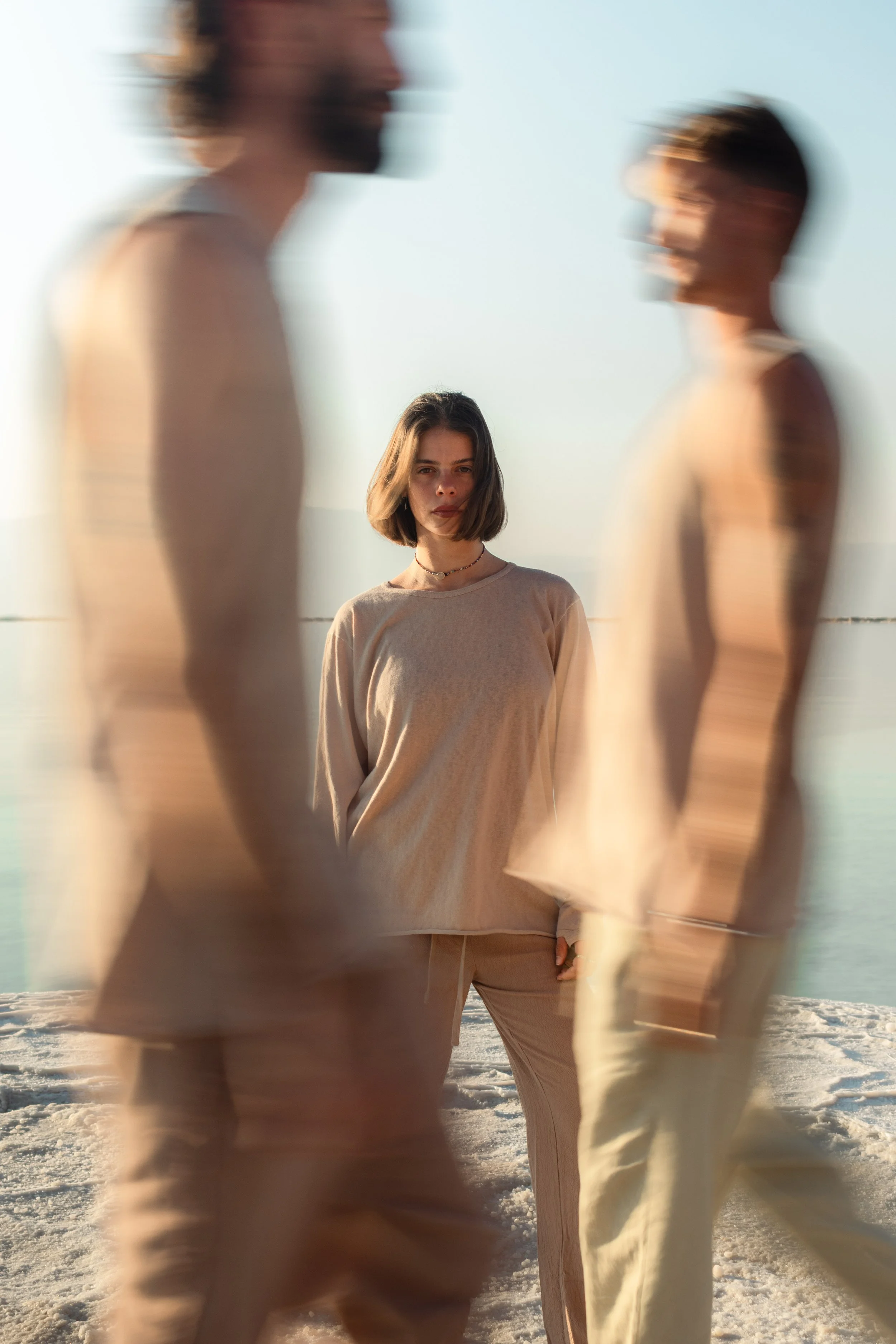 A woman standing on a beach with two people walking past her in the foreground, both blurry, with the sunlight illuminating her face and hair.