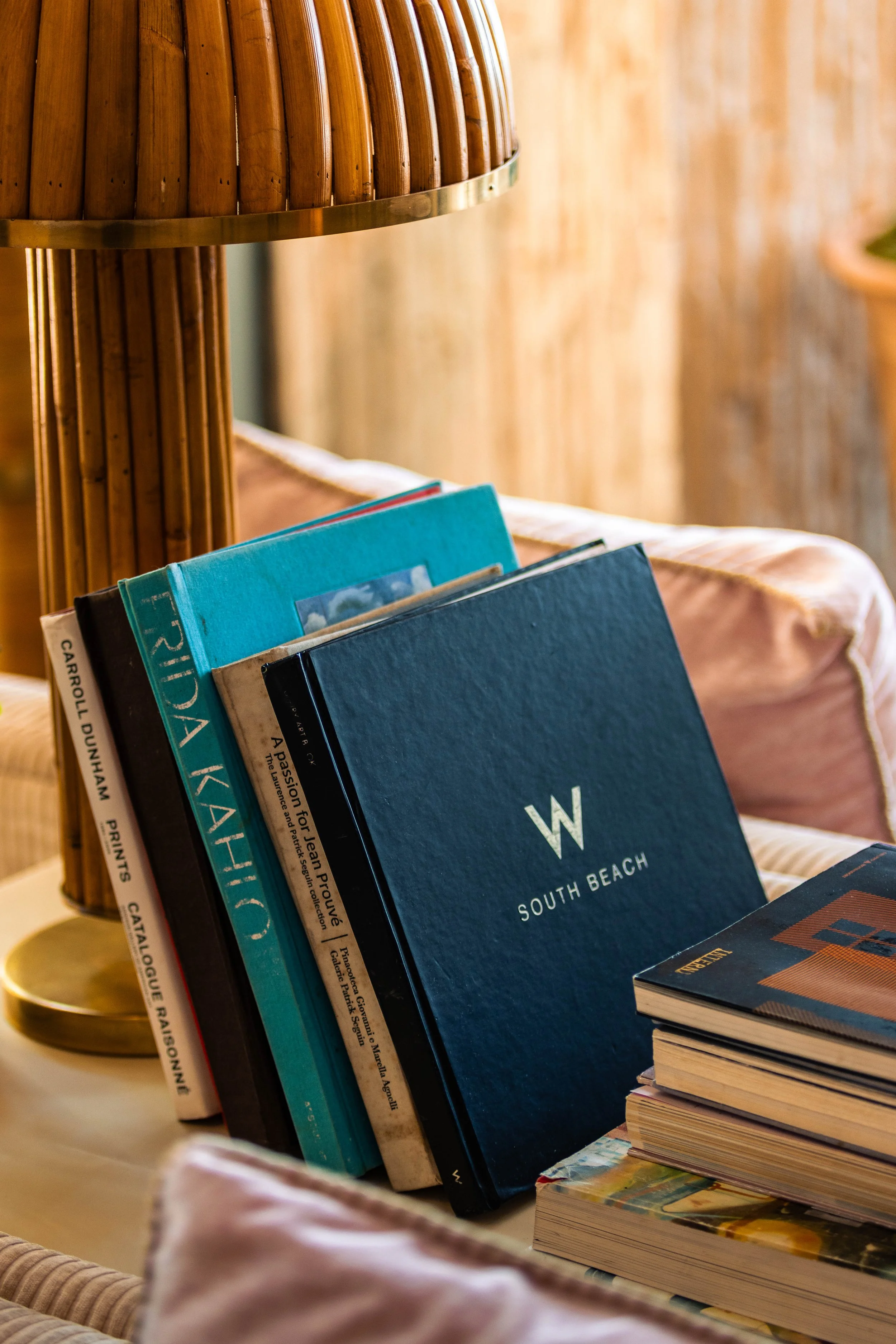 Books on a table in front of a bamboo lamp, with a light-colored couch and wooden wall in the background.