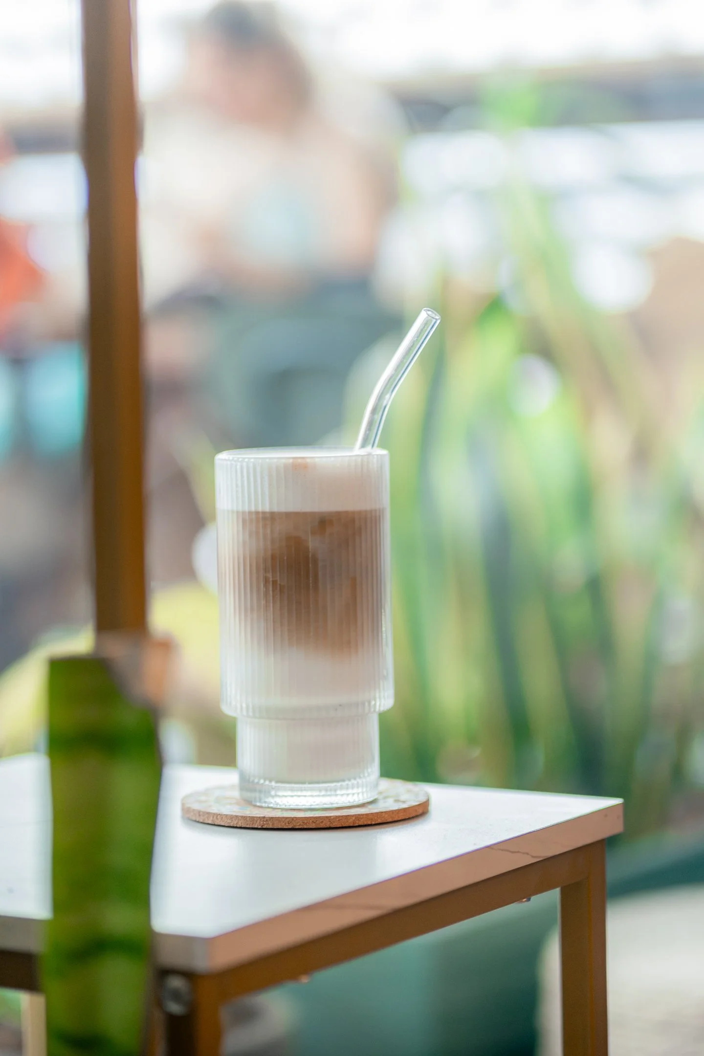 A tall glass of iced coffee with a ridged texture, topped with foam, and a straw, sitting on a wooden coaster on a small table in a cafe.