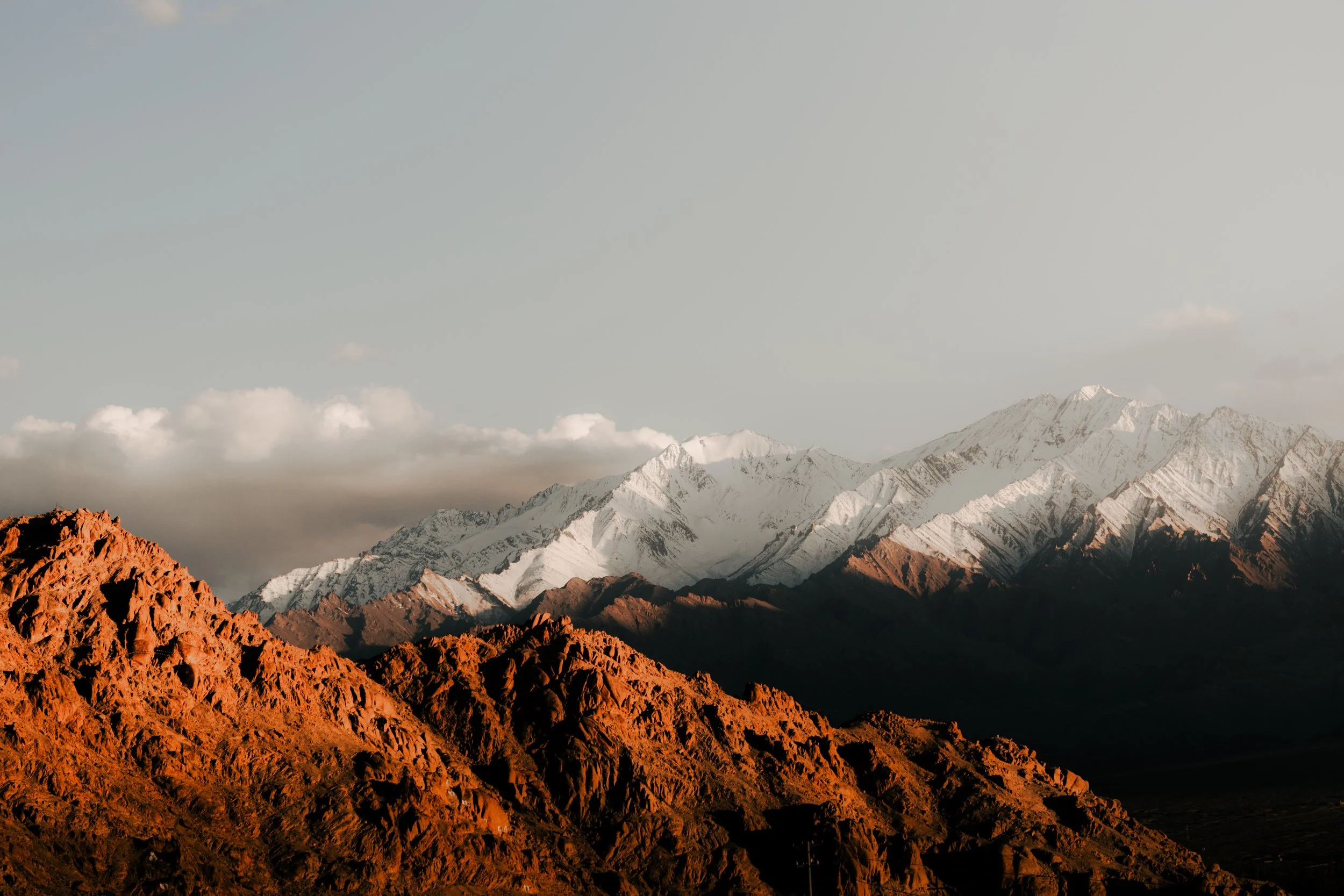 Mountain range with reddish-brown foreground rocks, snow-capped peaks in the background, and a partly cloudy sky.