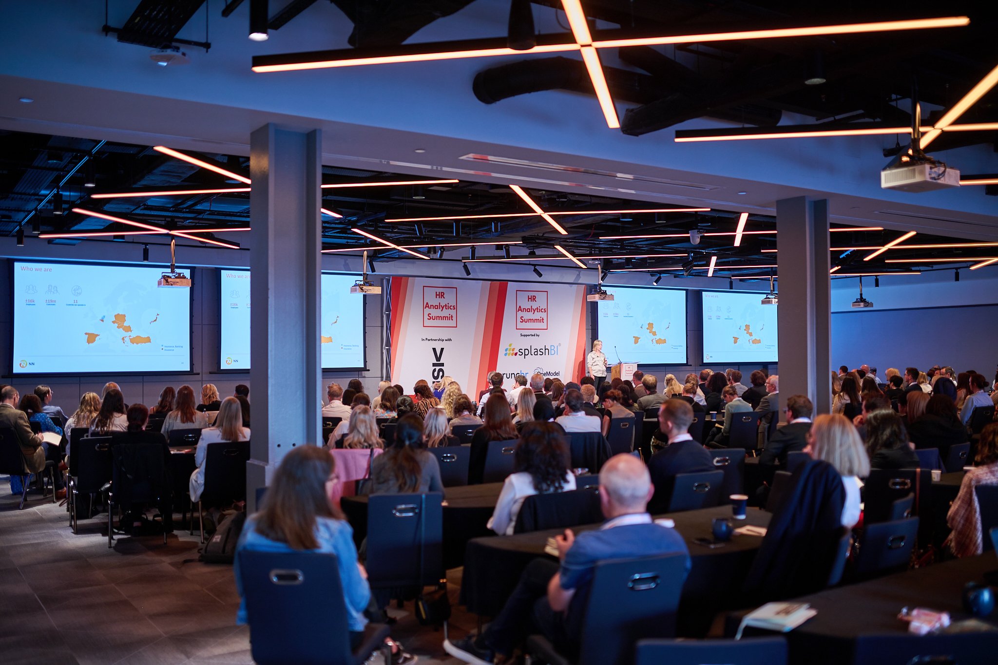 Conference attendees seated, facing a speaker at the HR Analytics Summit, with presentation screens displaying maps and text.
