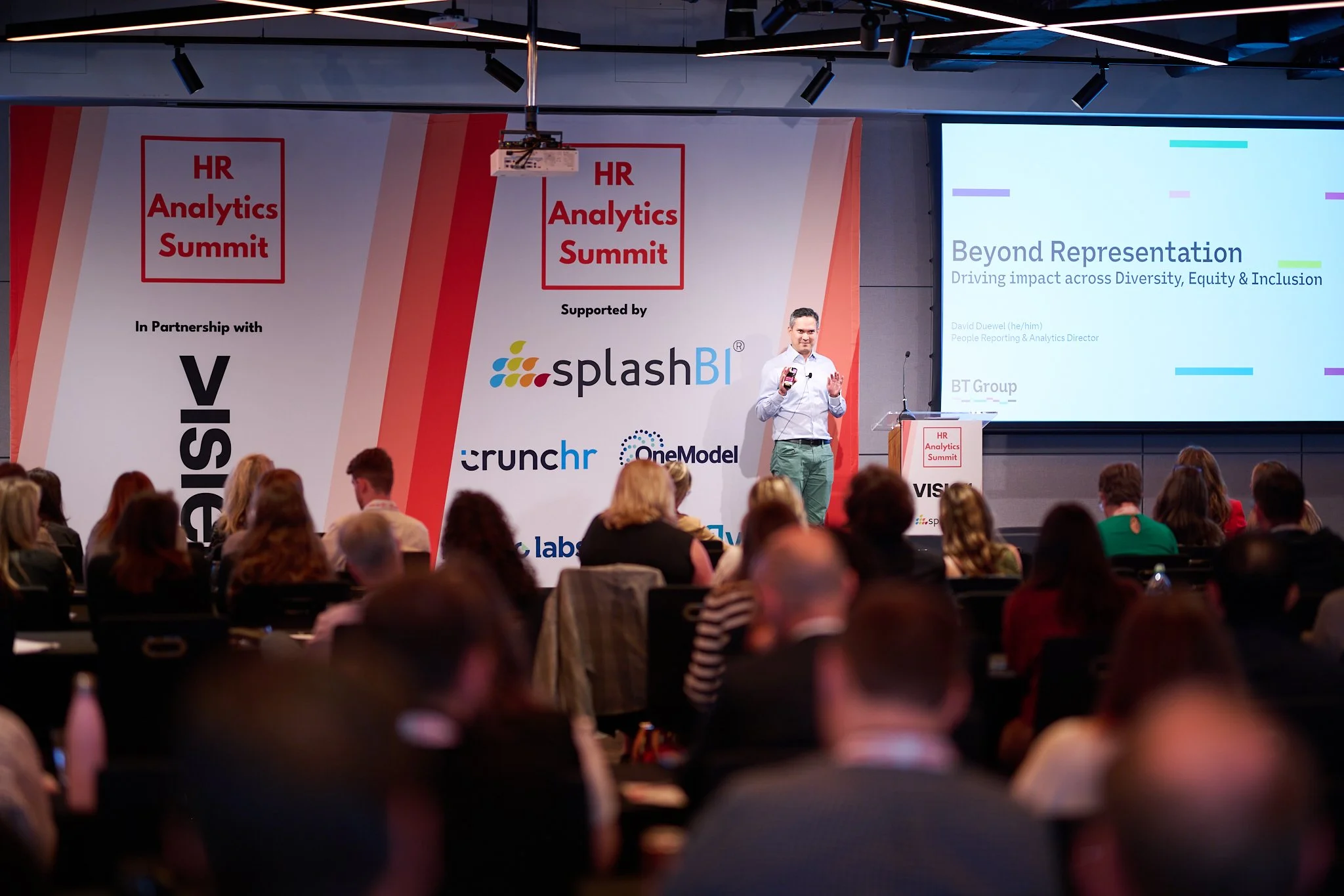 Speaker at HR Analytics Summit presenting on diversity, equity, and inclusion, with audience in attendance and event sponsors visible.