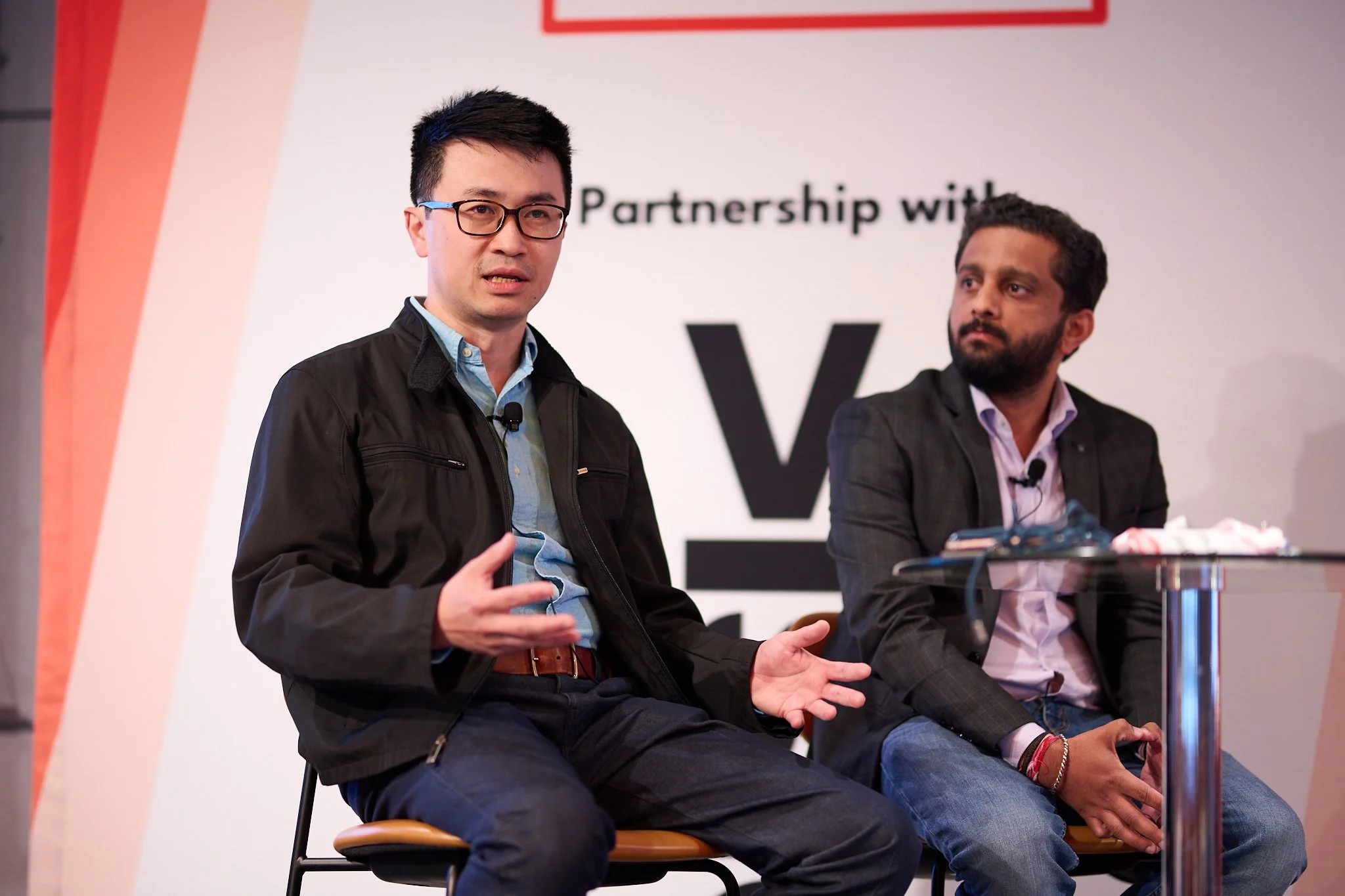 Two men speaking at a panel discussion, seated at a table with microphones, against a backdrop with some text partially visible.