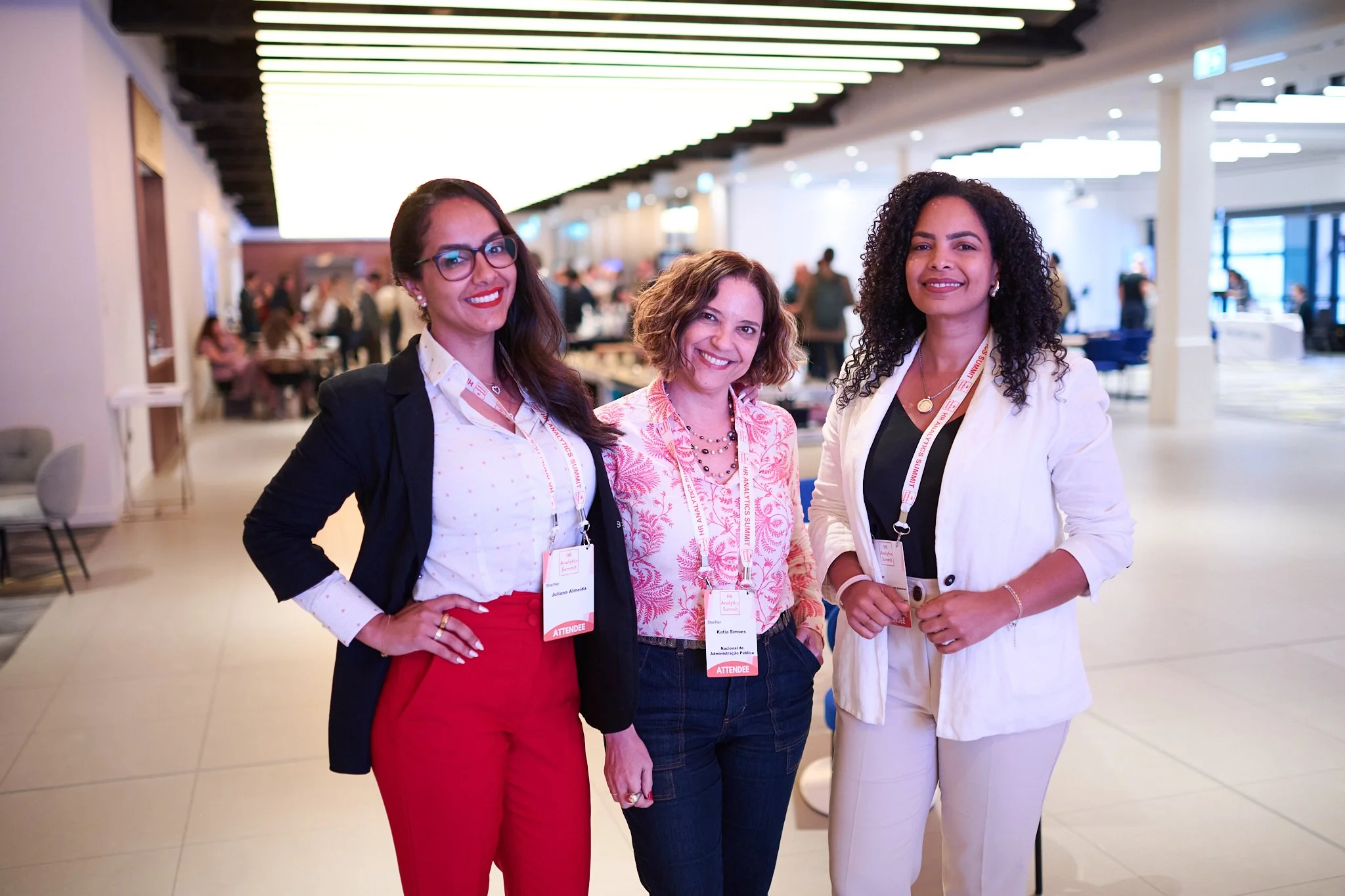 Three women at a conference wearing name badges and business attire, standing and smiling in a well-lit venue with people in the background.