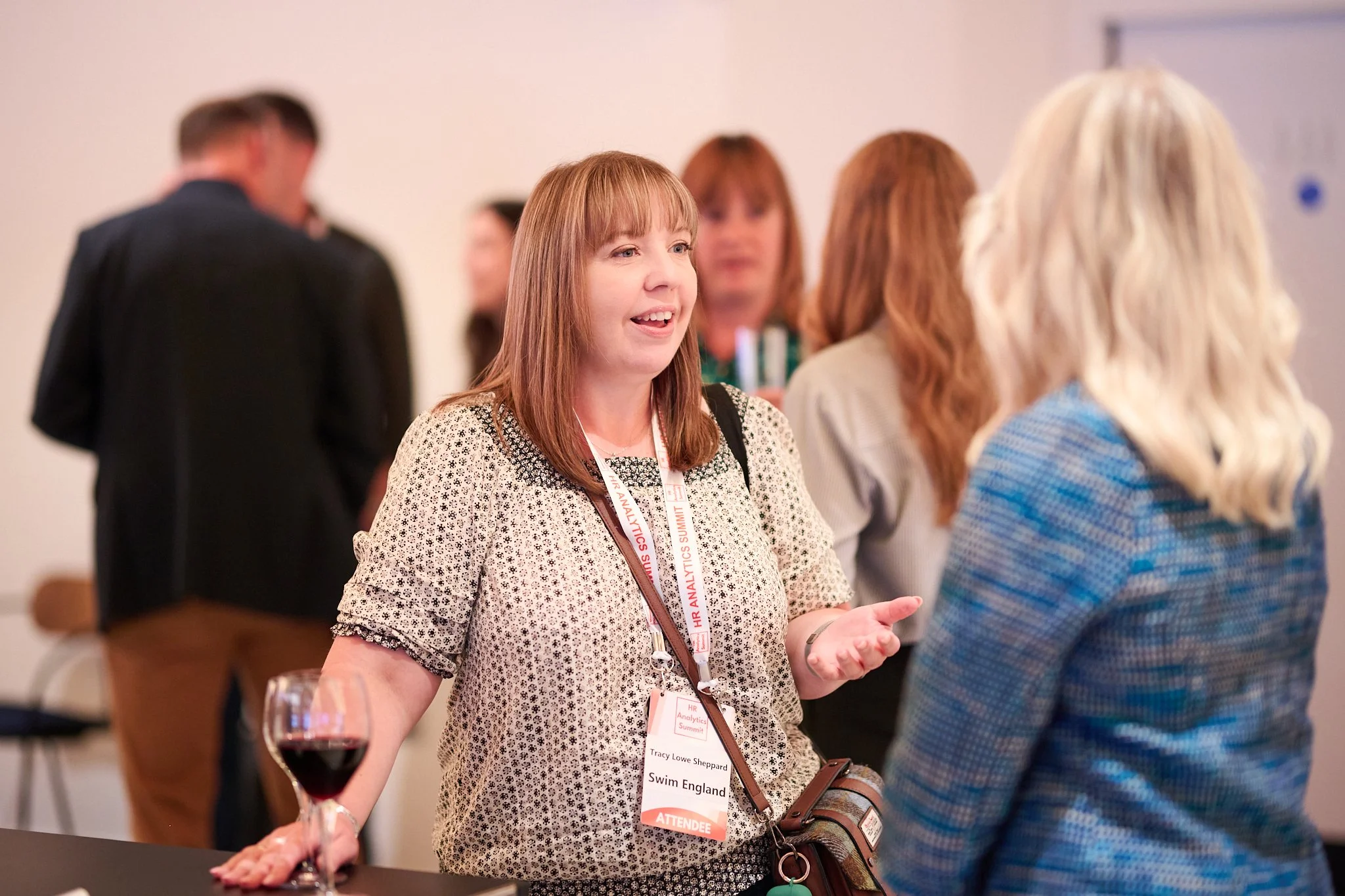 People networking at a professional event, woman in patterned blouse speaking, wearing a name badge, holding a glass of red wine.