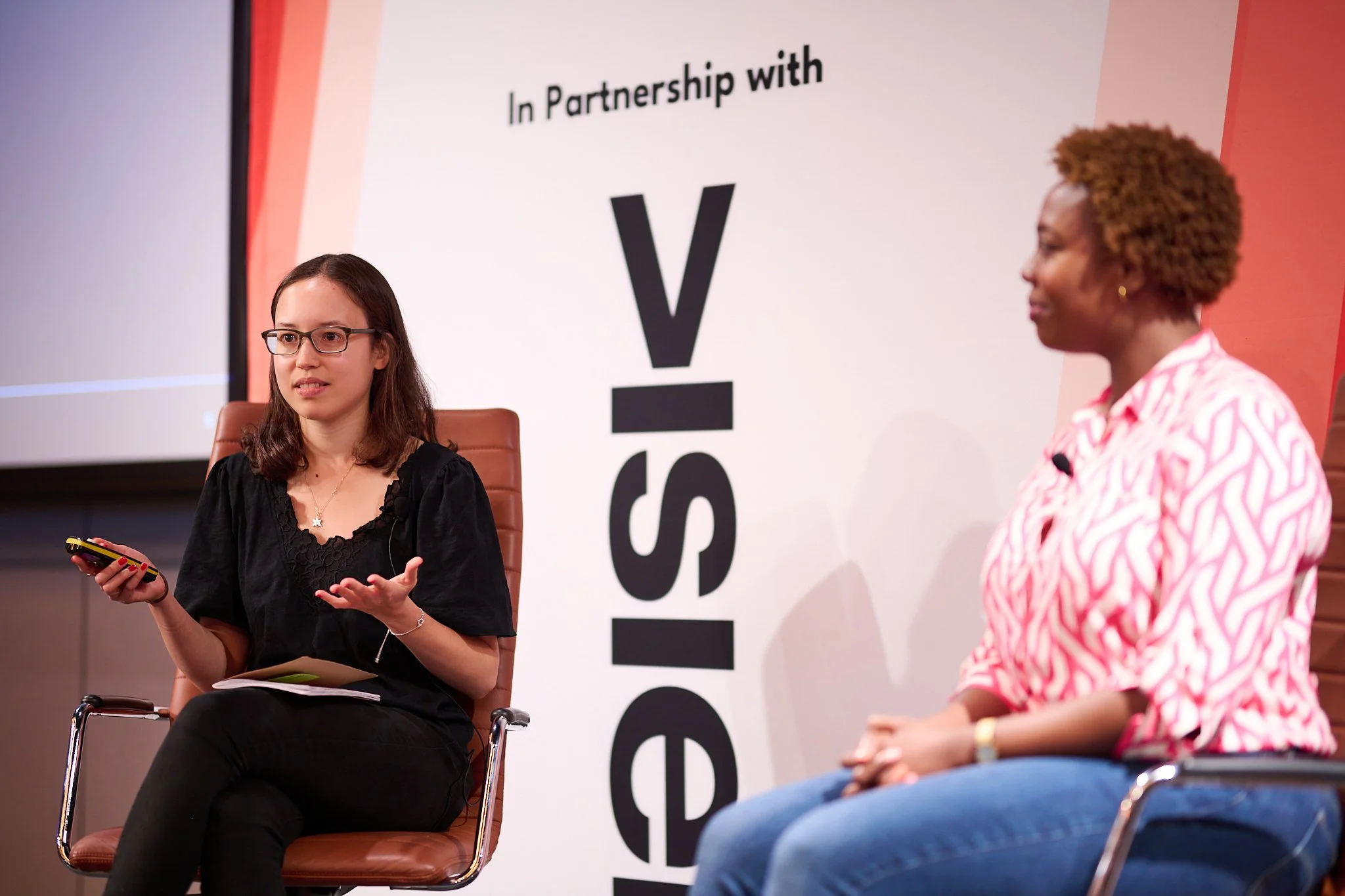 Two people seated on stage engaged in a discussion. One person is holding a smartphone and gesturing, while the other sits attentively. A backdrop displays partial text reading 'In Partnership with.'
