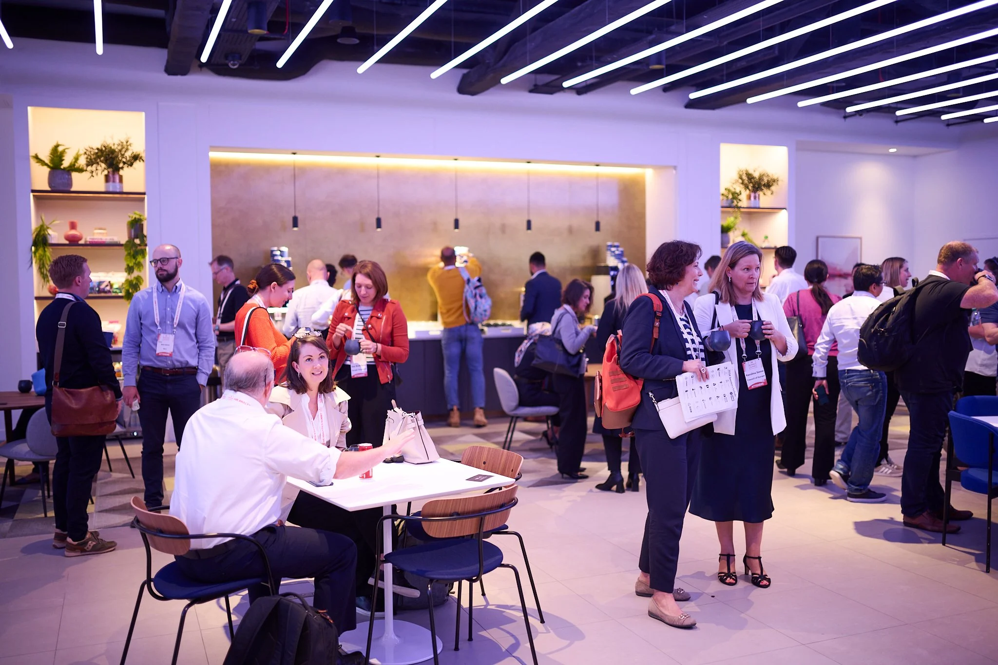 People networking at a conference event in a modern indoor space with tables, chairs, and plants.