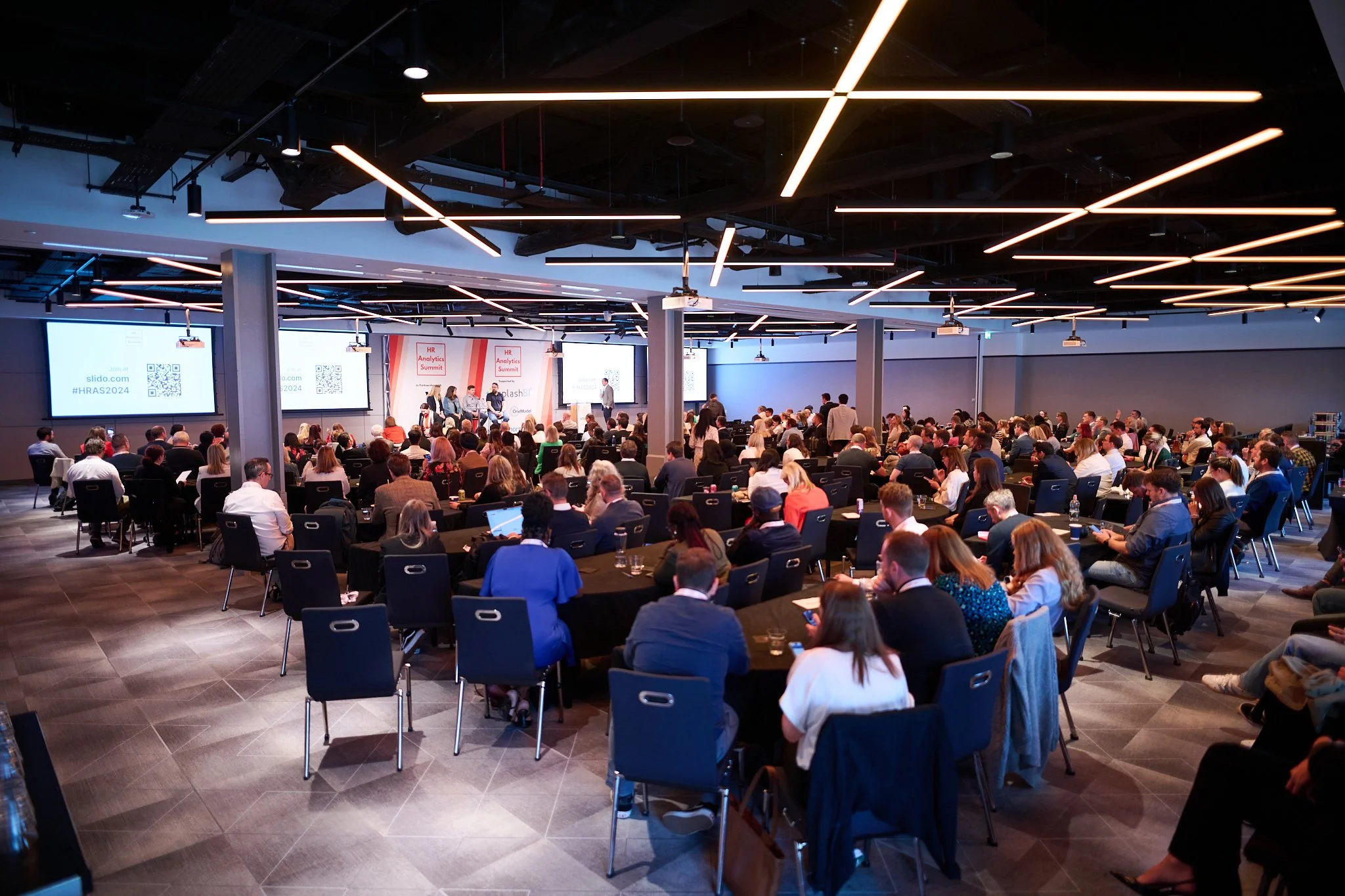 Large conference room filled with attendees watching a panel discussion on stage, with slides displayed on large screens.