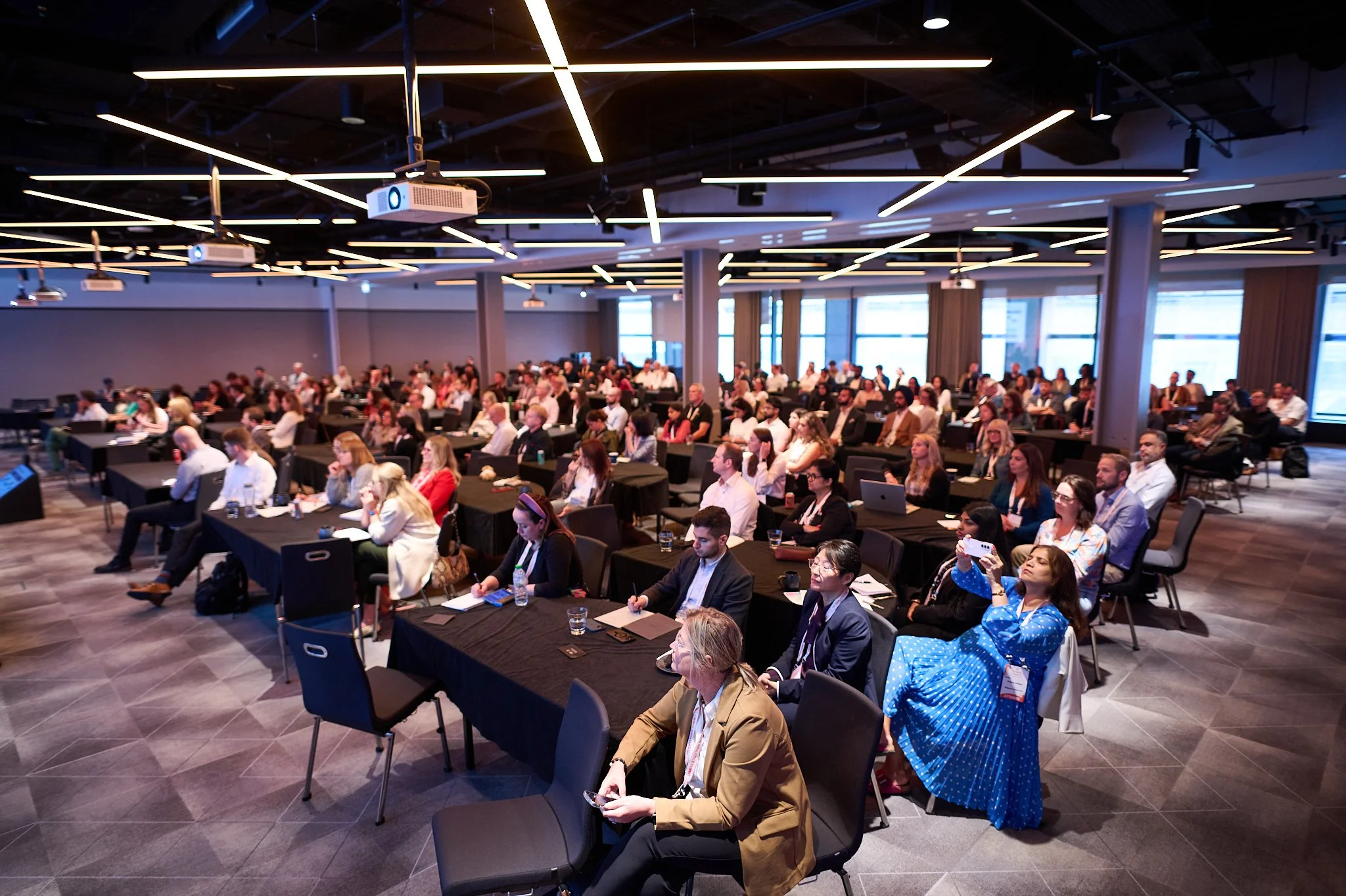 Audience attending a large conference with people seated at tables in a modern meeting room, multiple projectors visible on the ceiling.
