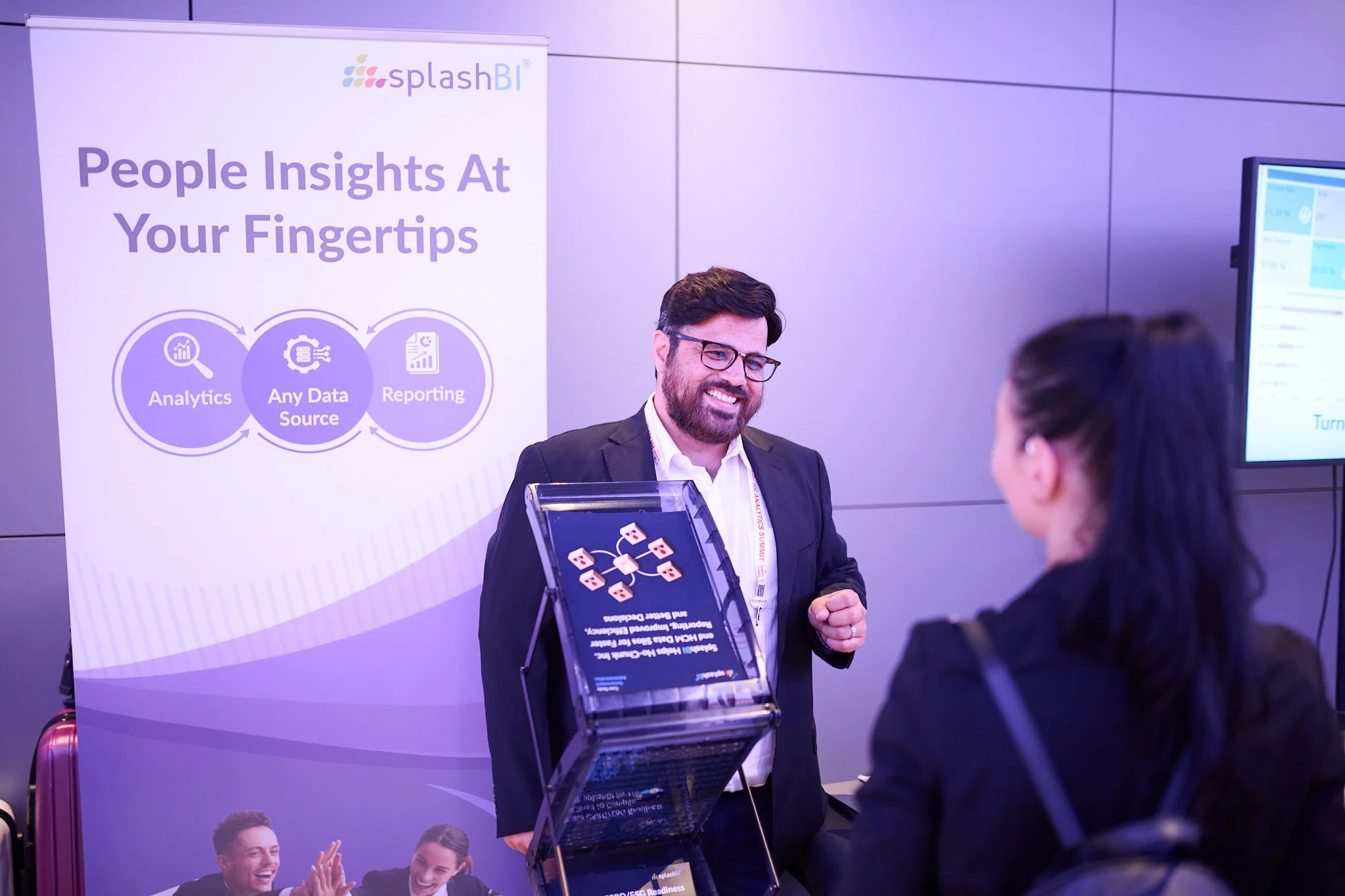 A man is smiling while standing at a booth with a display for "splashBI." The booth features a banner with the text "People Insights At Your Fingertips" and icons for analytics, data sources, and reporting. A woman is engaged in conversation with him