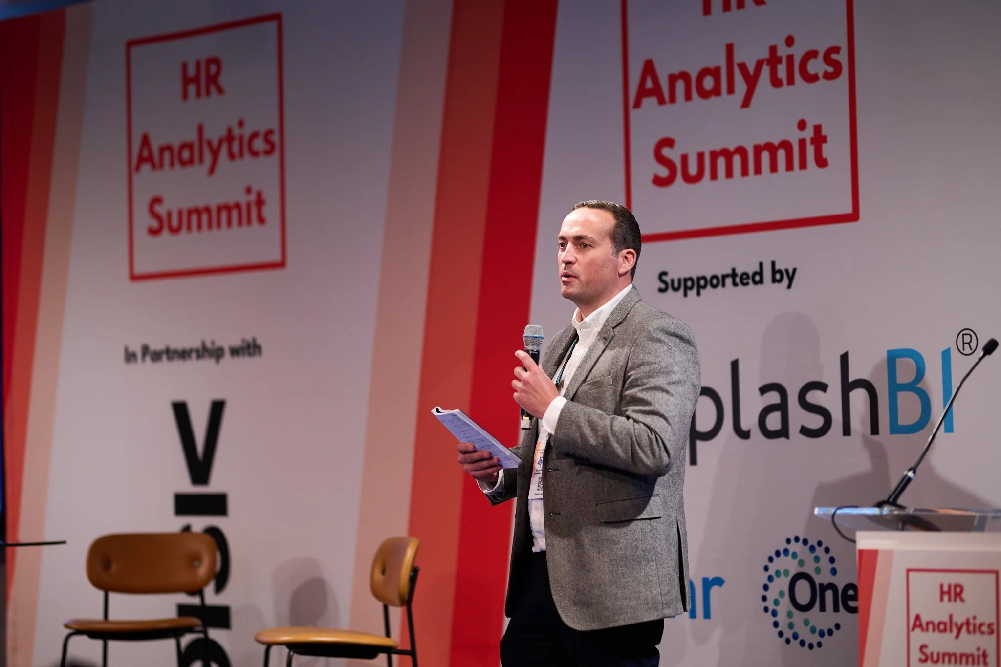 Speaker holding a microphone at HR Analytics Summit with banner and chairs in background.