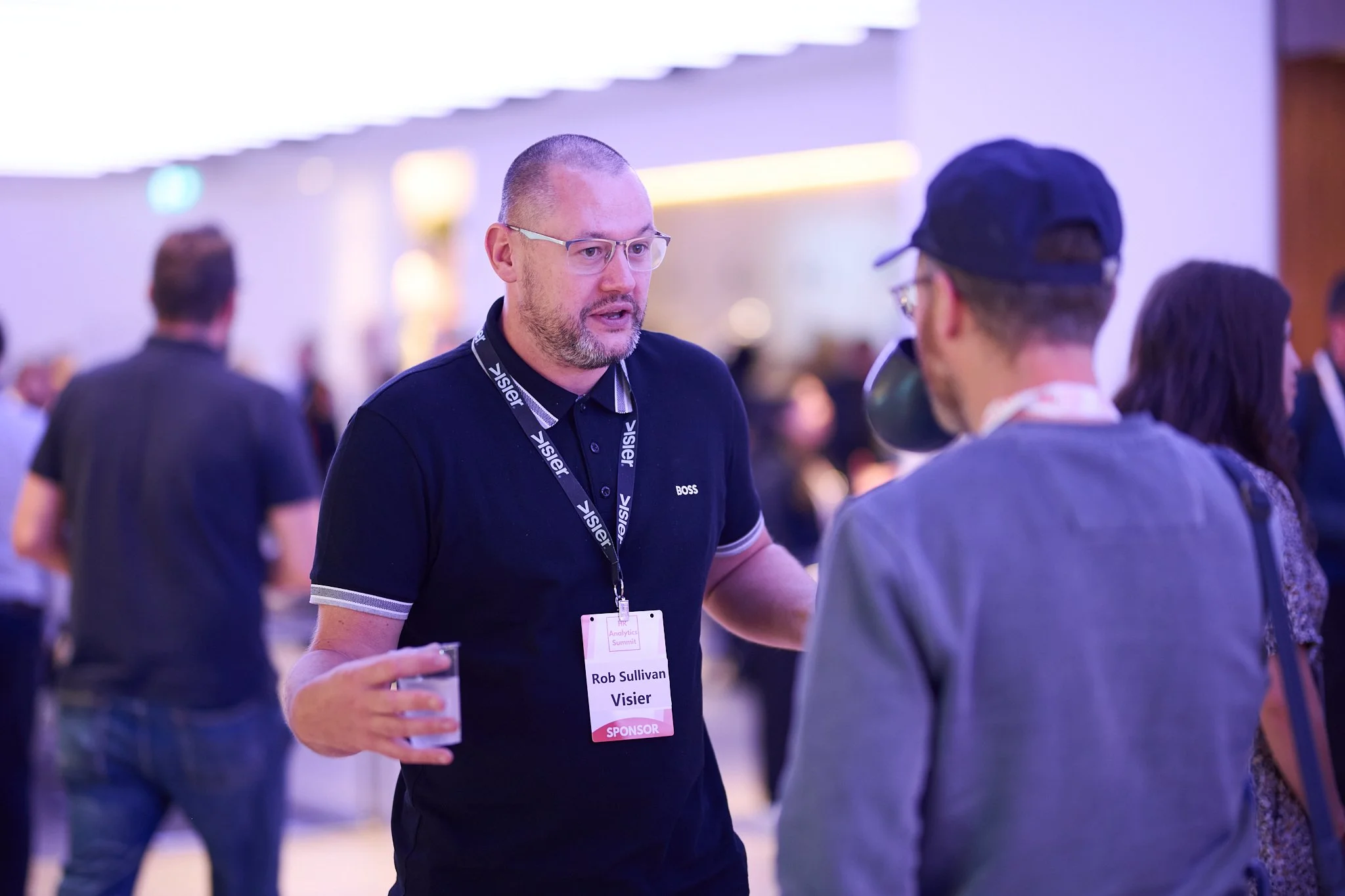 Two men conversing at a conference event, one wearing a black shirt and glasses with an event badge. The setting is a busy, indoor space with other attendees in the background.