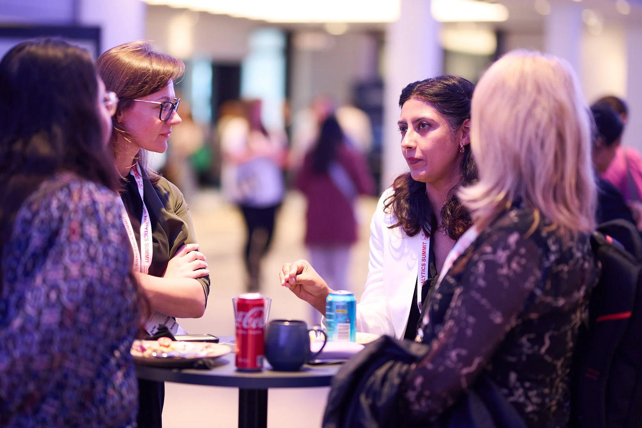 Group of people having a conversation at a table with drinks, including a can of Coca-Cola and a can of Pepsi, in a conference setting. Participants are wearing lanyards and discussing intently.