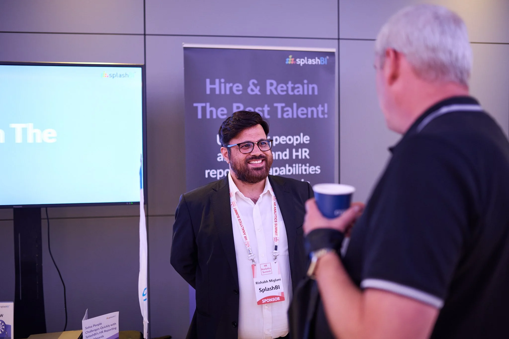 Two men talking at a business event, one wearing a name tag that says 'SplashBI Sponsor.' There's a banner behind them promoting employee and HR capabilities.