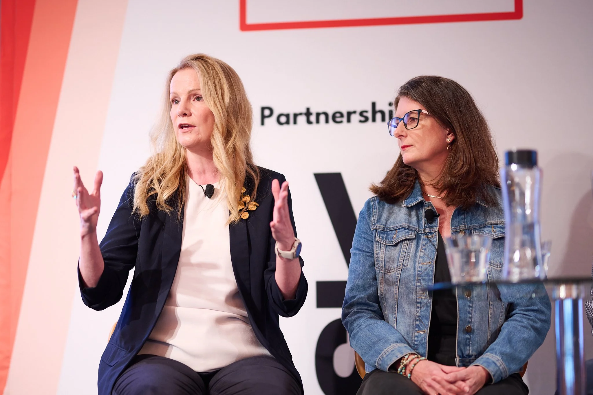 Two women sitting, engaged in a discussion at a conference, with a backdrop featuring a logo and the word 'Partnership.' One woman is gesturing while speaking, and the other is listening attentively. A table with glasses and a bottle is in the foregr