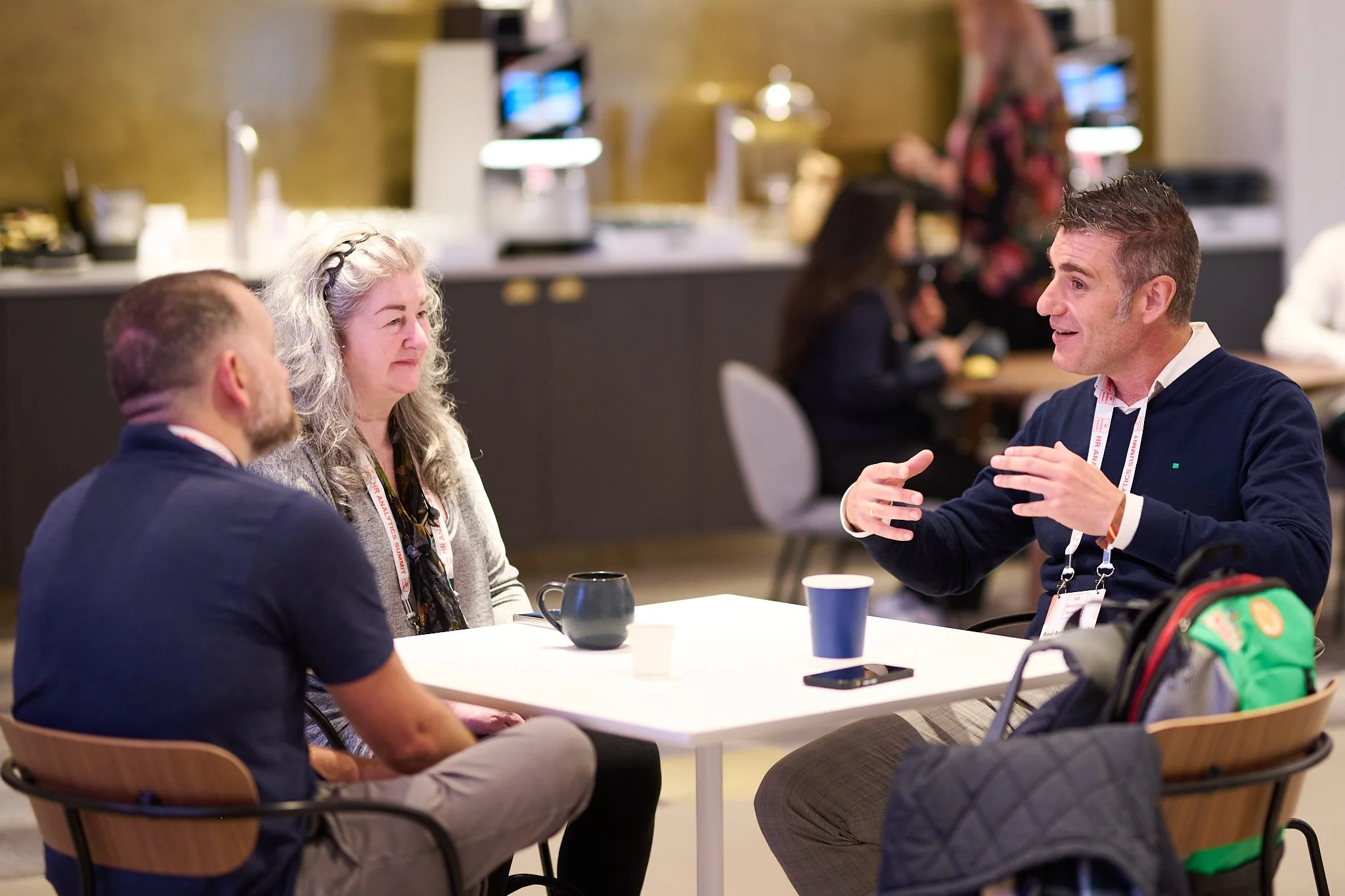Three people engaged in a conversation at a table in a casual setting, with coffee cups on the table and other people in the background.