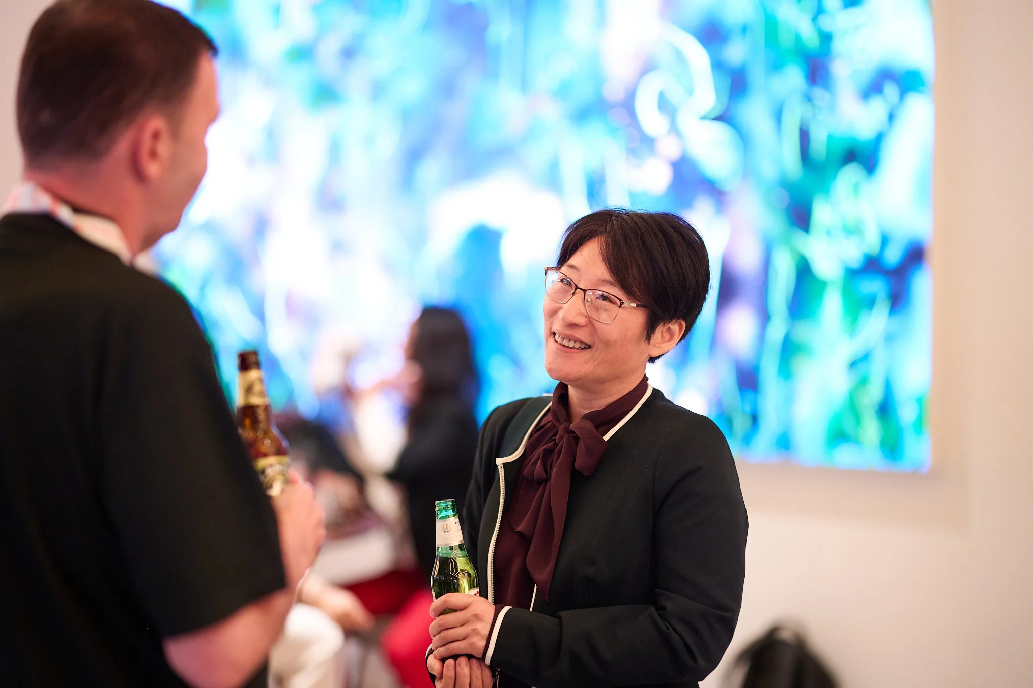 Two people chatting at a social event, holding bottles, with a colorful abstract background.