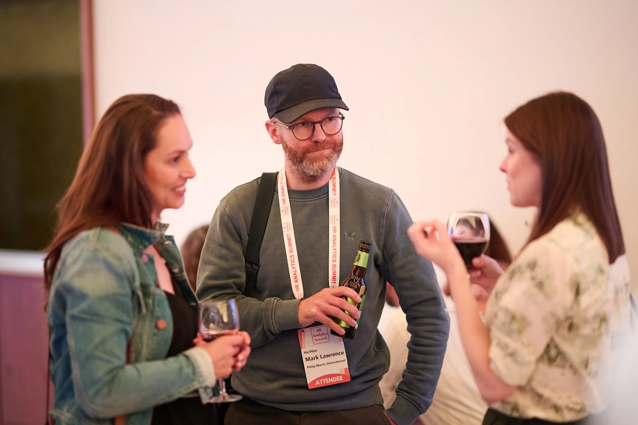 Three people at a social event, two women holding wine glasses and a man with a beer bottle and lanyard.
