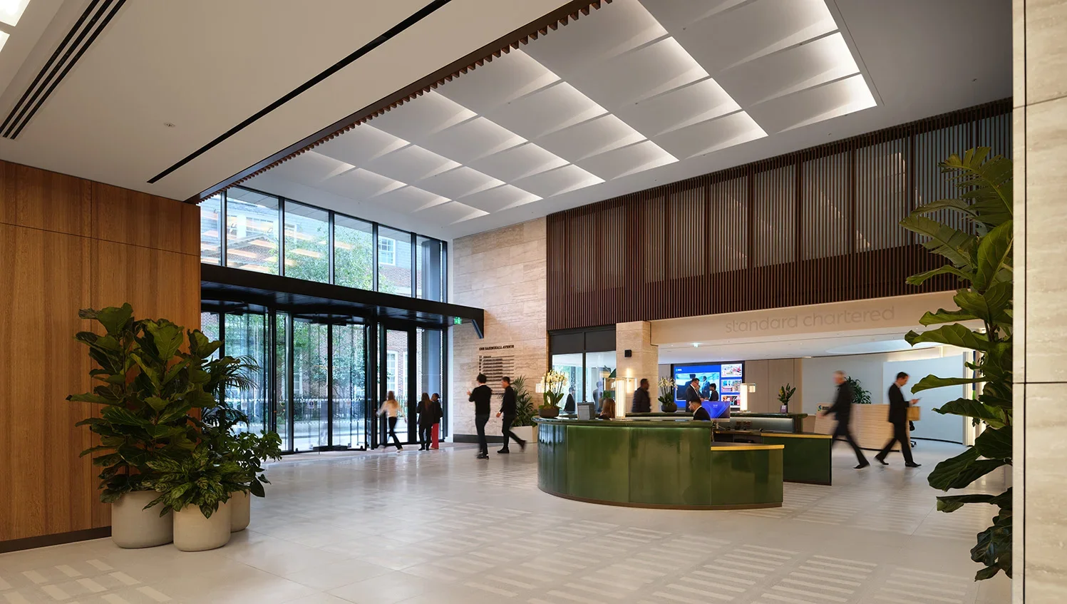 Hotel lobby with large glass entrance, wooden wall paneling, indoor plants, a green reception desk, and people walking through.