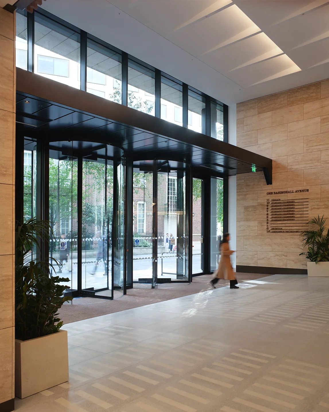 Modern building lobby with glass revolving door, indoor plants, and directory signage on beige stone wall.
