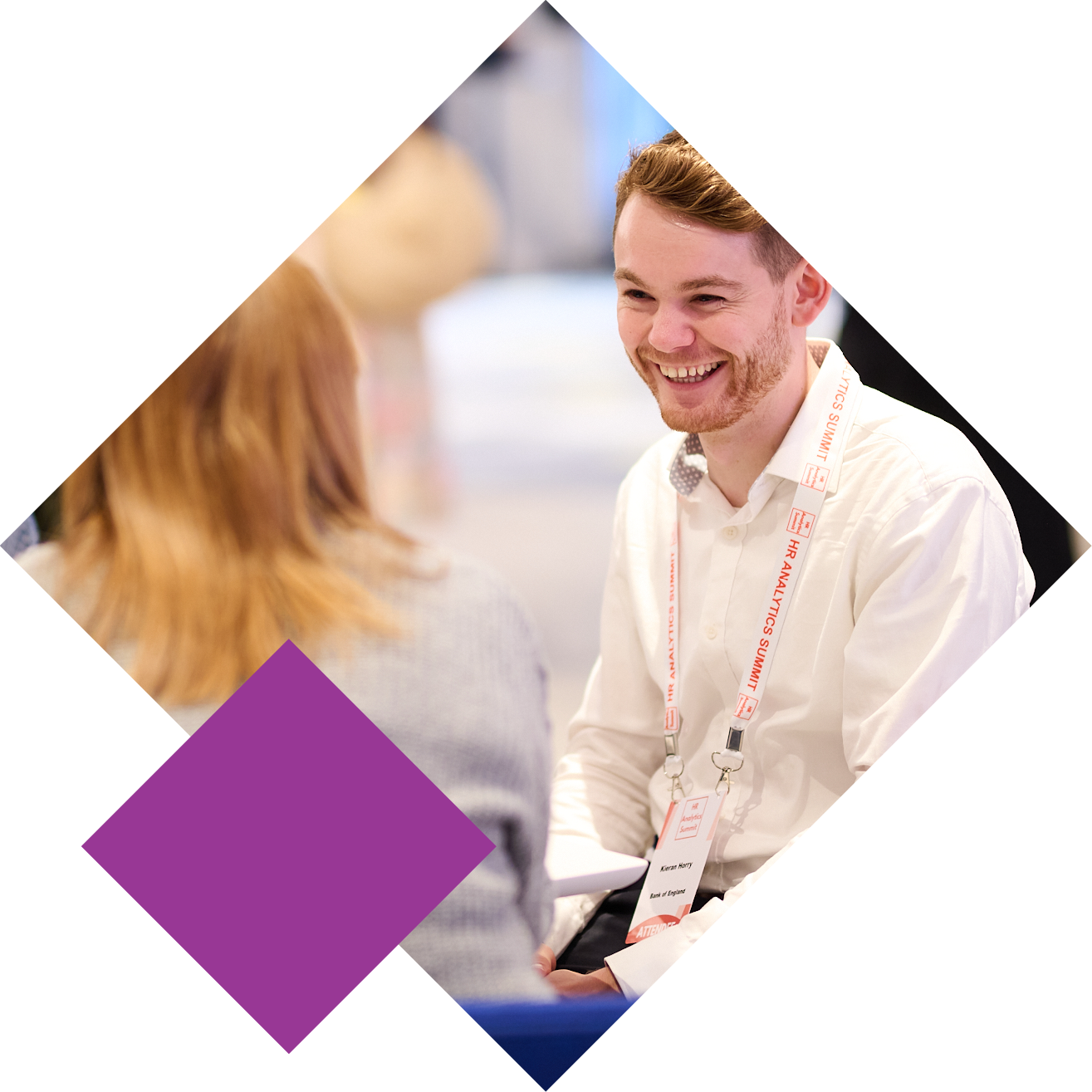 Two people talking at a professional event, one facing the camera, wearing a lanyard and smiling.
