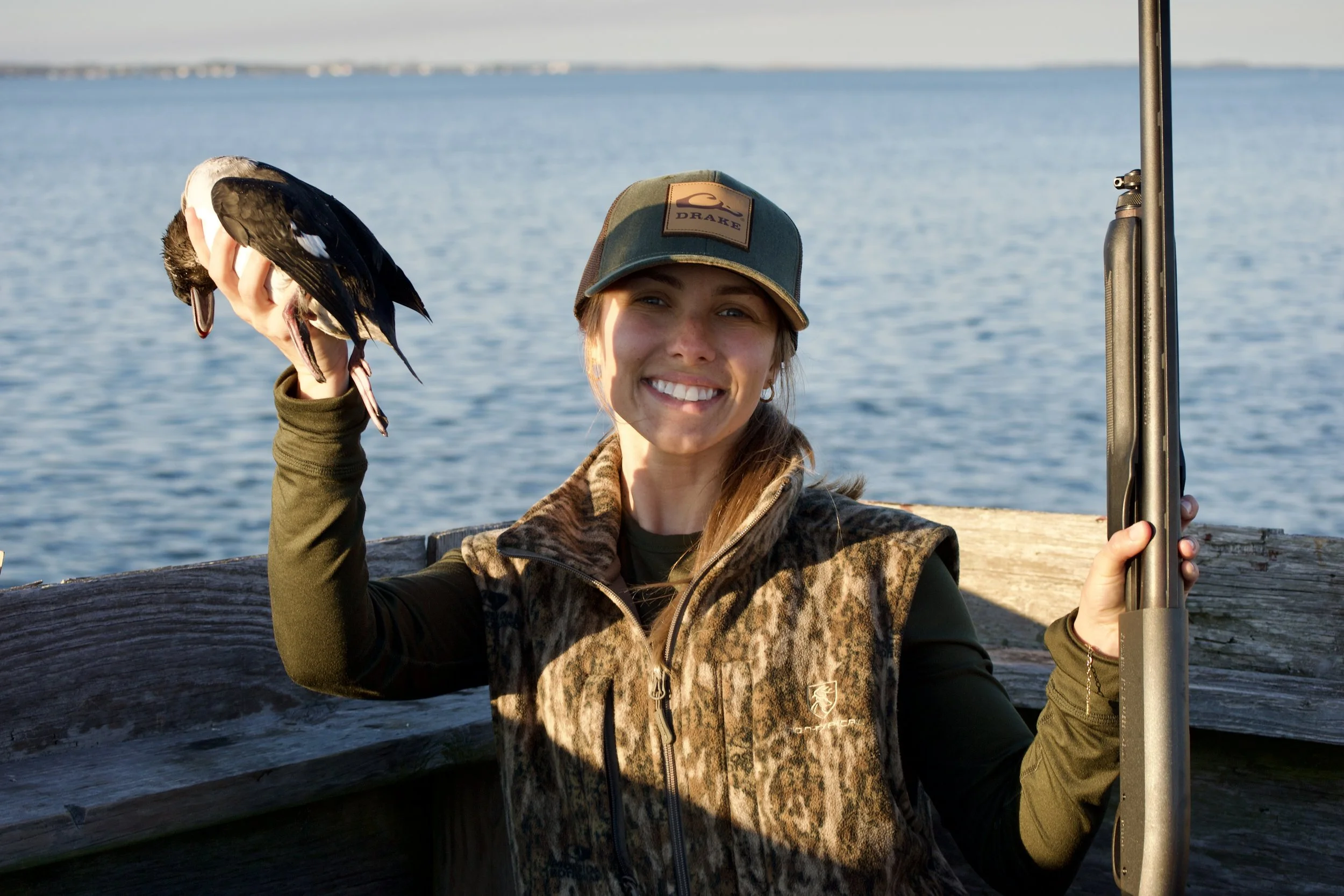A woman smiling and holding a bird next to her face, standing on a dock with water in the background, wearing a cap and outdoor gear.