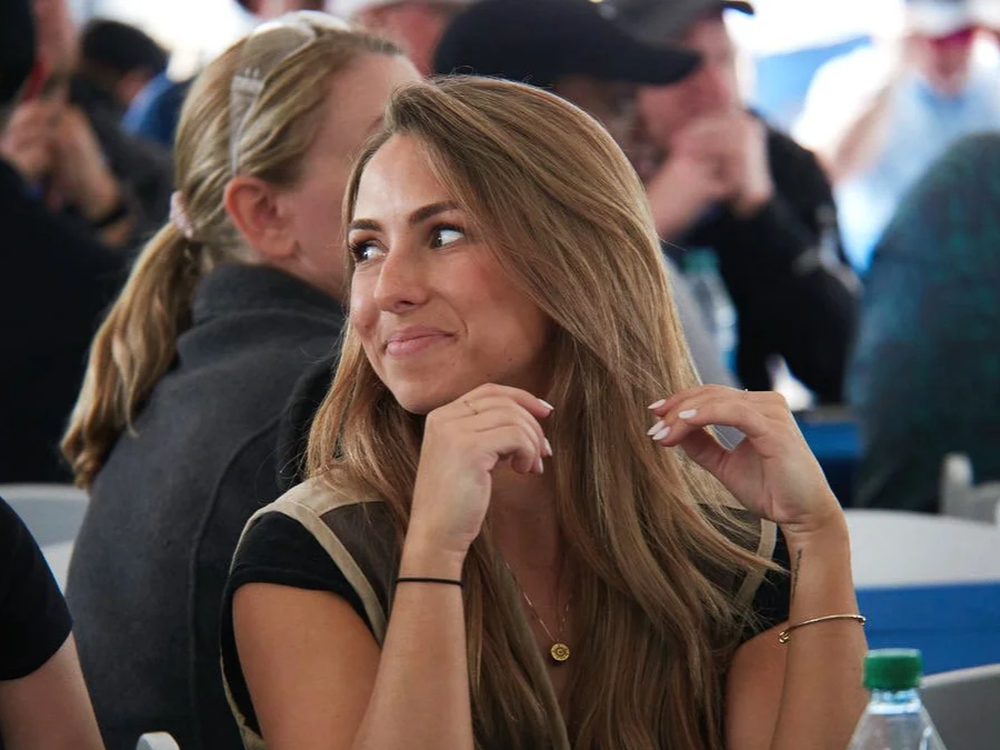 A young woman with long, light brown hair sitting at a table at a social gathering, smiling and looking to her right, with other people visible in the background.