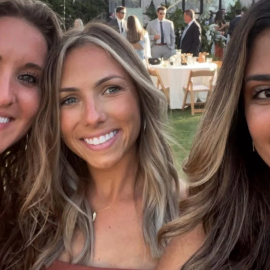 Three women smiling and posing for a photo at an outdoor event with a table and other people in the background.