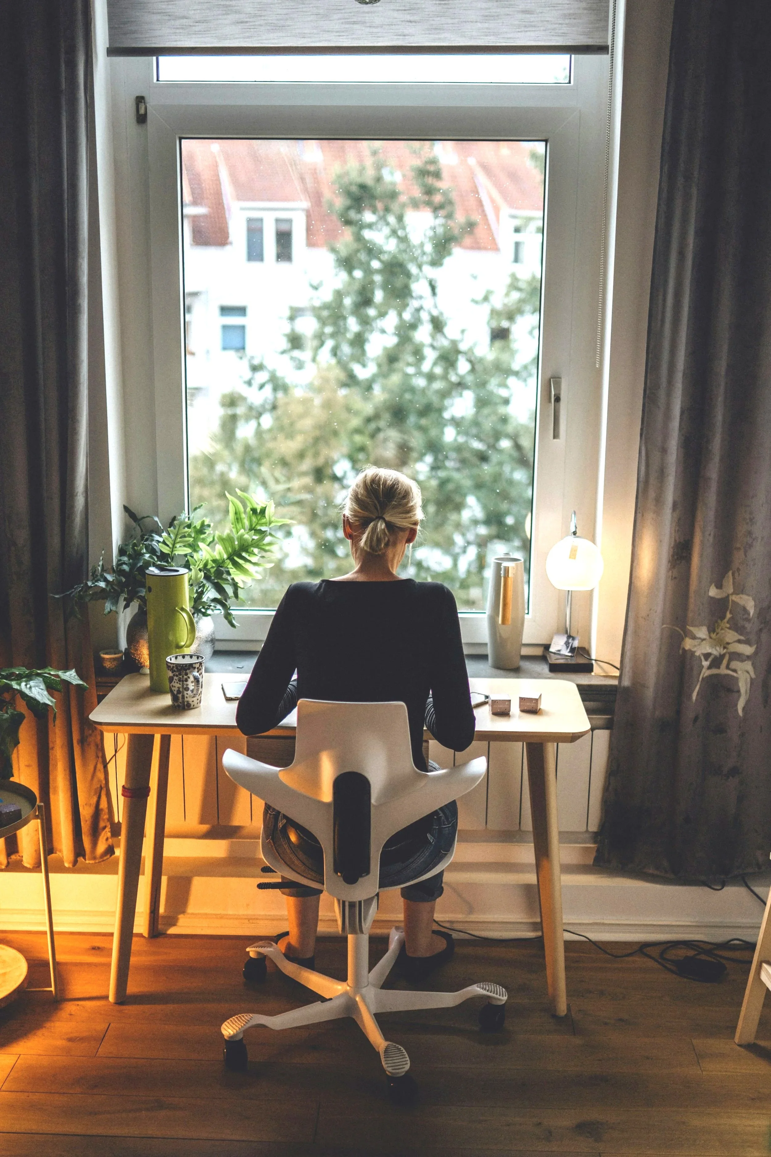 Paperwings & Co. LLC., Tori Jackson. Corporate business travel services. A woman with blonde hair tied back, sitting at a desk by a large window with a view of trees and buildings, in a cozy, well-lit home office with plants and a lamp.