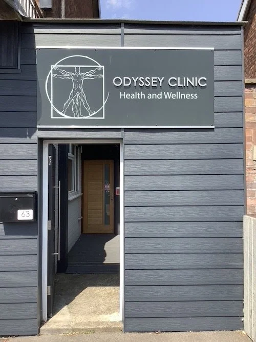 Exterior view of Odyssey Clinic, Leamington Spa, with a sign displaying a Vitruvian Man illustration. The entrance has an open door leading inside, and the building's exterior is clad in horizontal grey panels.