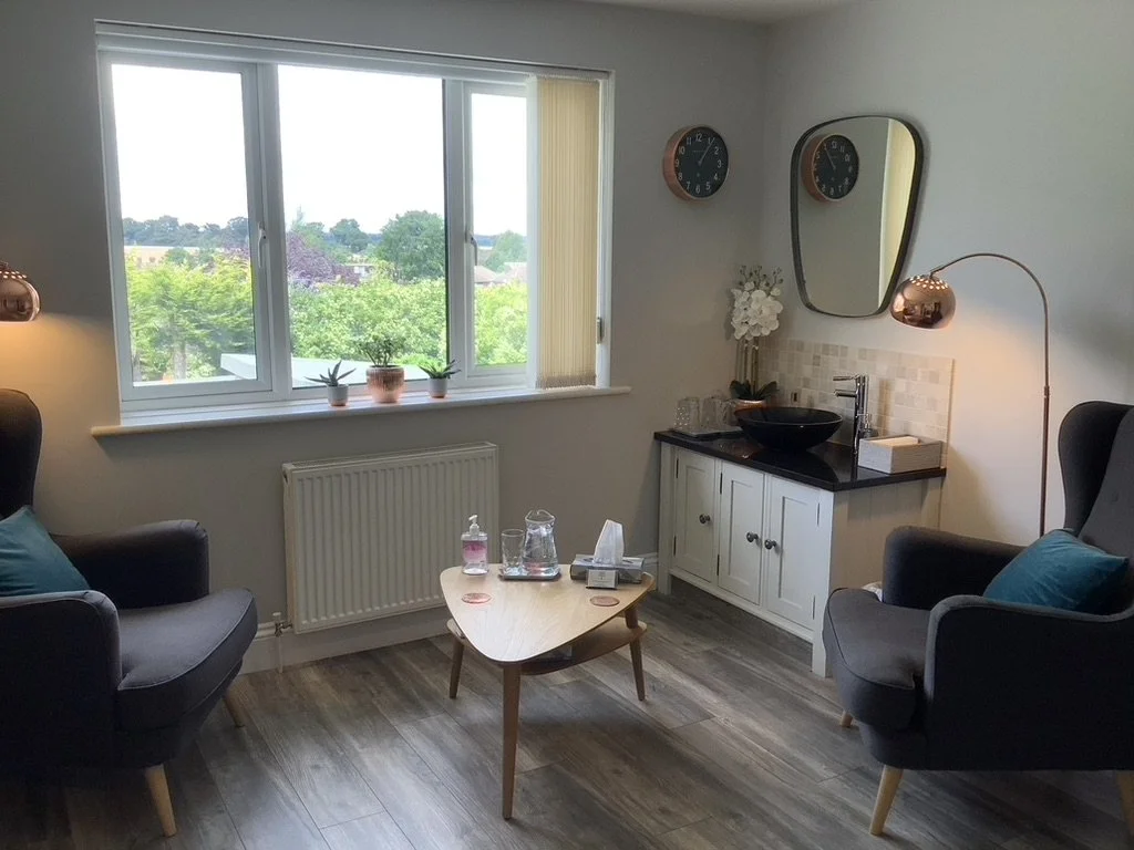 Interior view of Clear Insight Counselling therapy room, with two grey armchairs, a small wooden coffee table, and a black sink with a clock and mirror hanging above. A large window has a view of green trees outside and floor lamps provide lighting.
