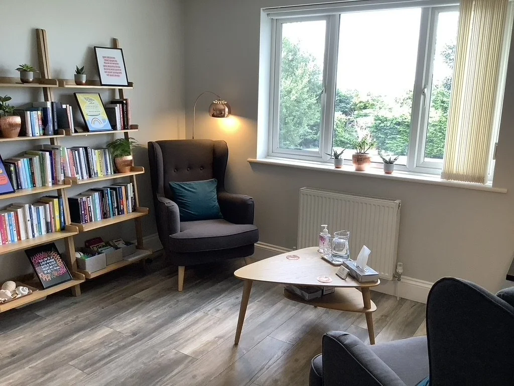 Interior view of Clear Insight Counselling therapy room, with two grey armchairs, a wooden bookshelf filled with books and potted plants, a small wooden coffee table and a window with a view of green trees outside. A floor lamp provides lighting.