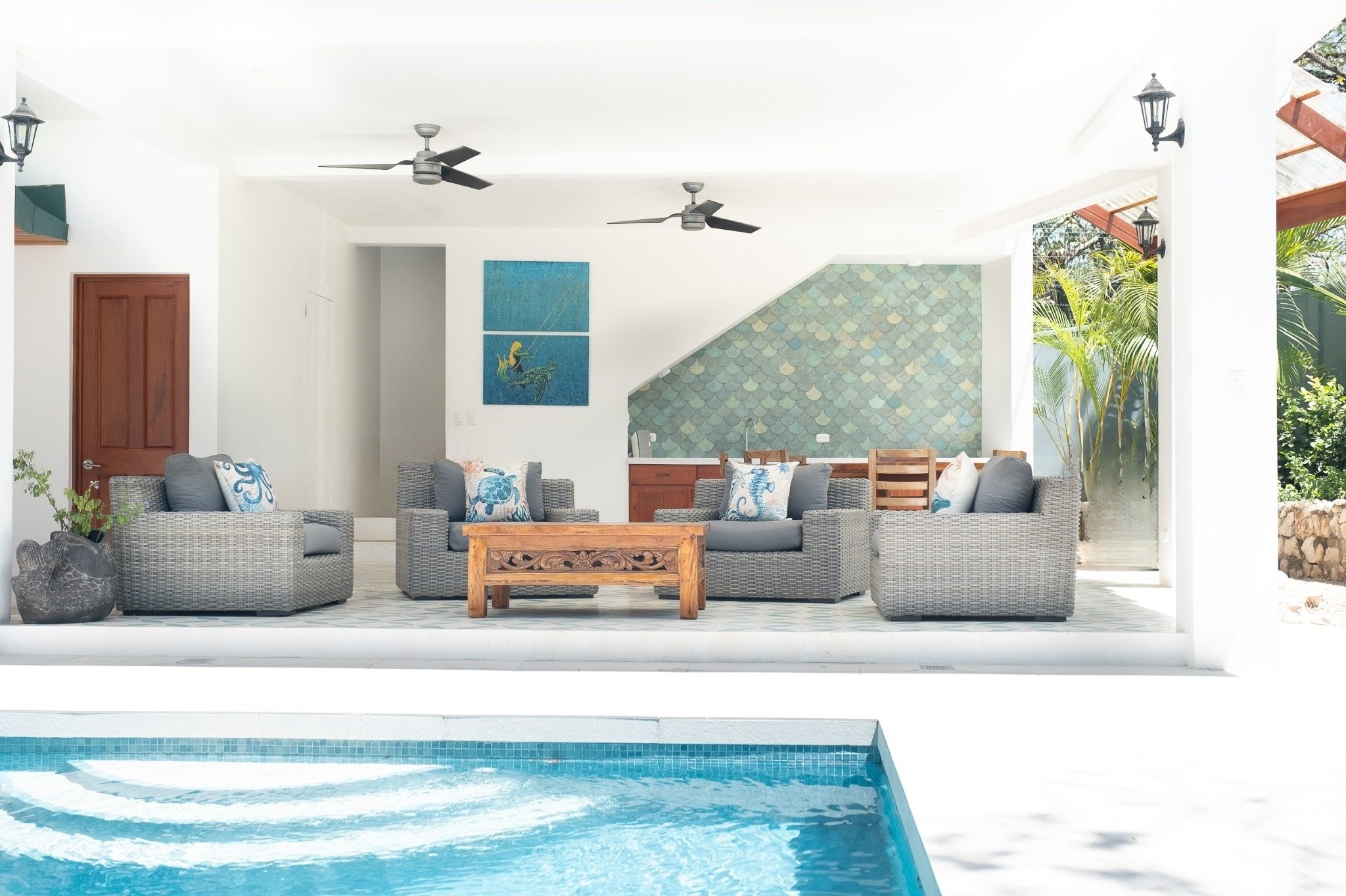 Outdoor pool area with four wicker sofas with grey cushions, blue and white pillows, a wooden coffee table, and a dining area with wooden chairs. The space features ceiling fans, wall-mounted lanterns, and tropical plants, with exterior walls painted