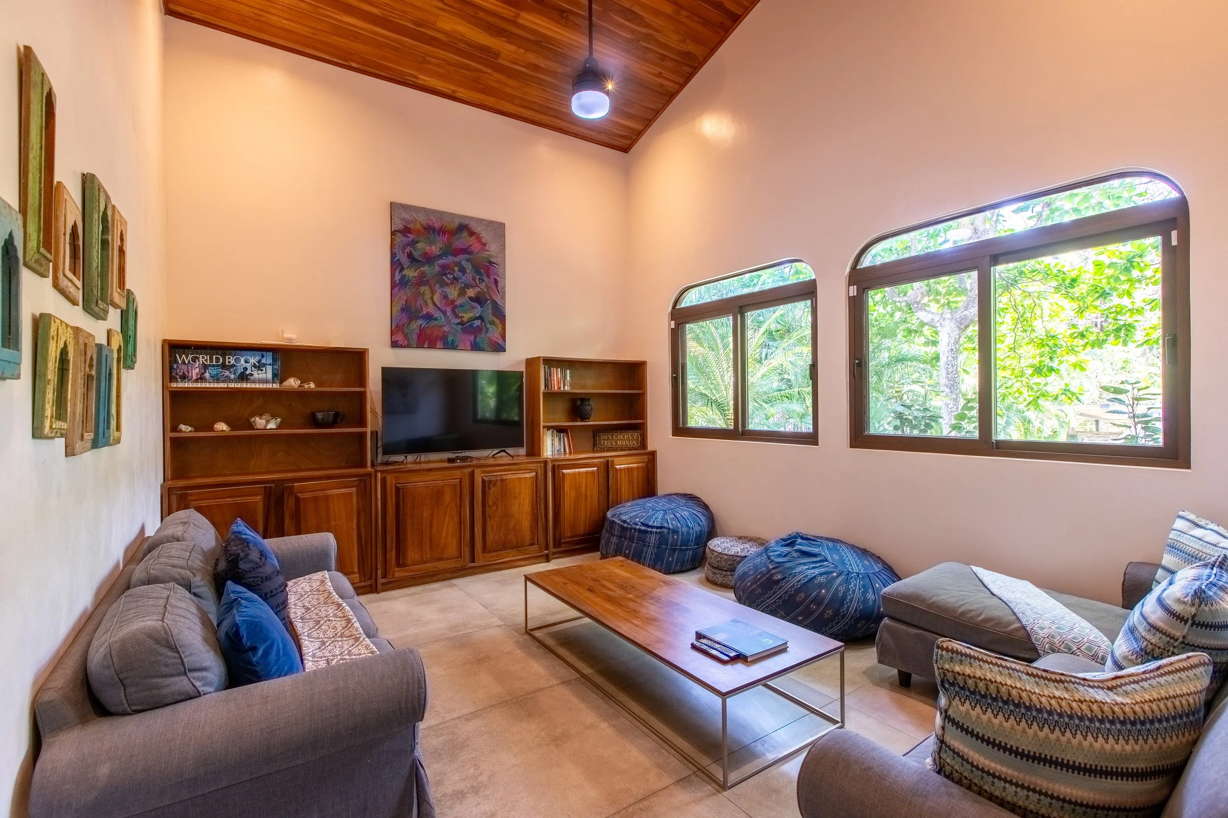 Living room with beige walls, wooden ceiling, windows with greenery outside, gray sofa with blue and patterned pillows, wooden coffee table, TV on wooden cabinet, colorful lion painting on wall, blue ottomans, armchair with patterned pillow.