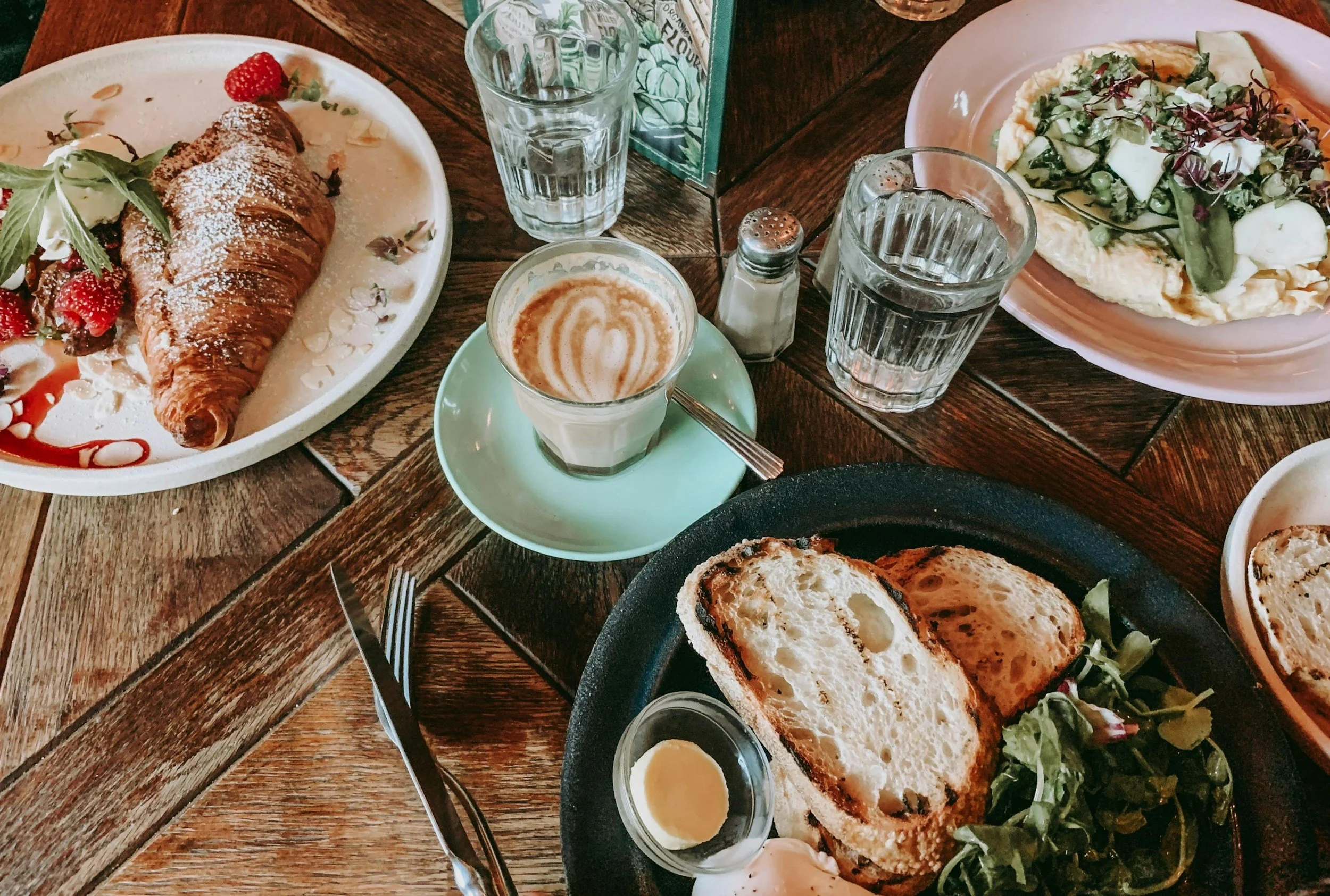 A wooden table with various breakfast foods including a croissant with strawberries on a white plate, a dish with eggs and greens, a toasted bread with butter, and a cup of coffee with latte art, along with glasses of water.