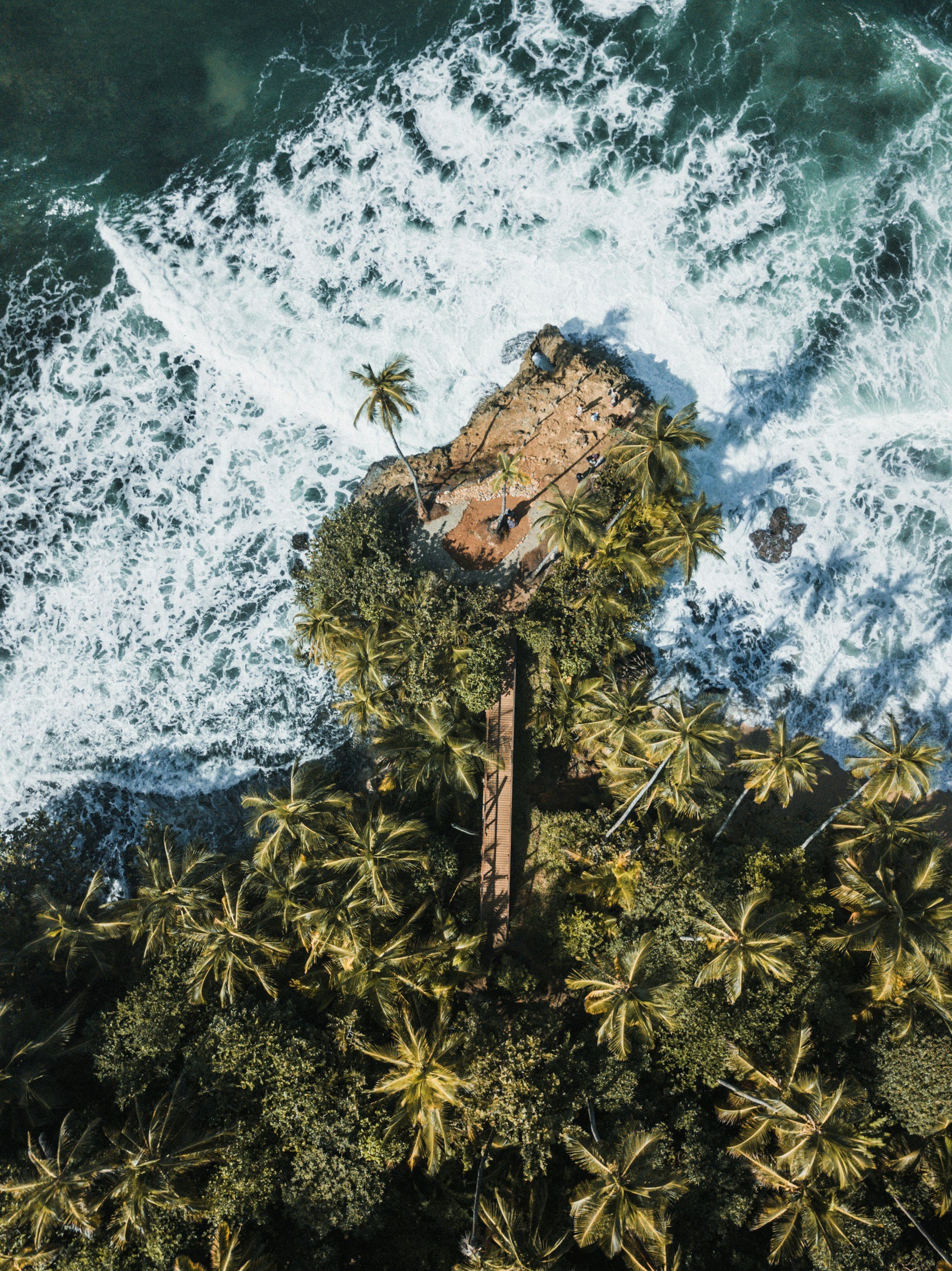 Aerial view of a small island with palm trees surrounded by ocean waves, with a pier extending from the island into the water.
