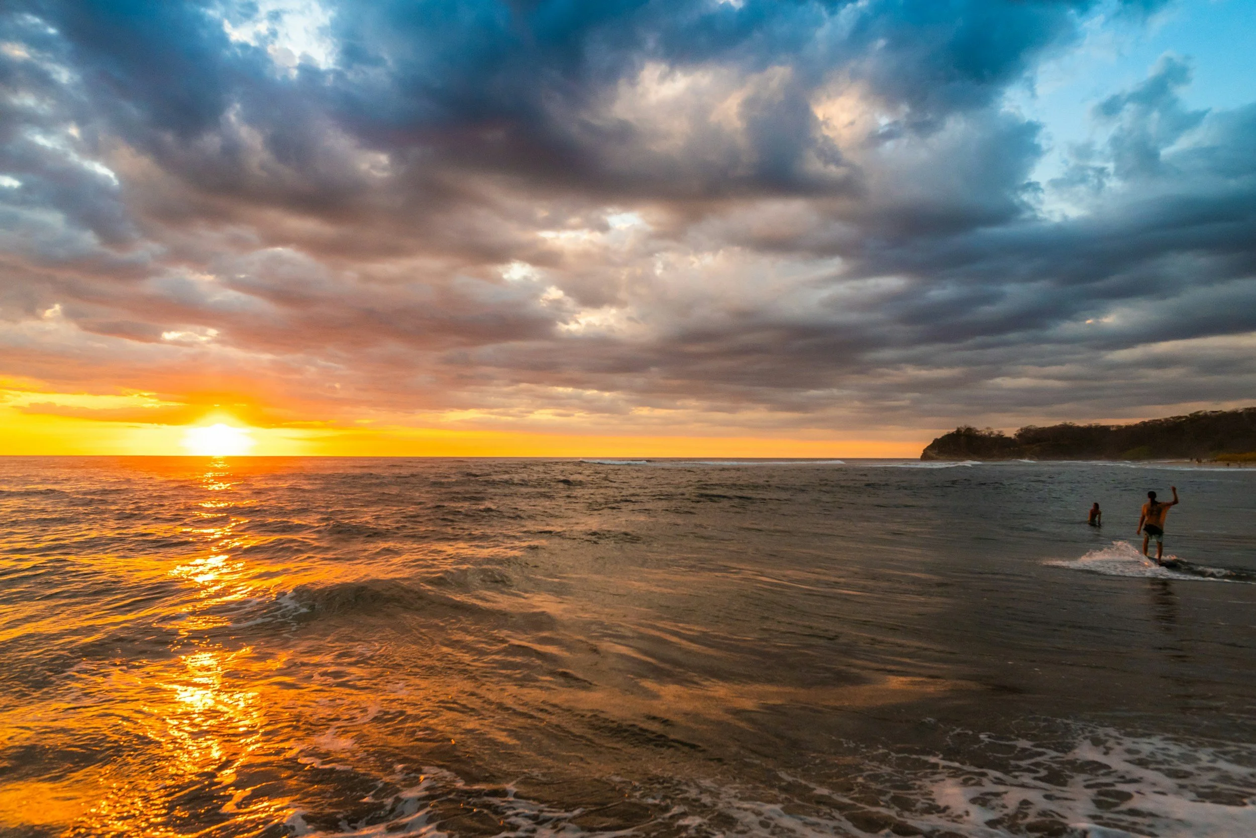 Sunset over the ocean with a cloudy sky and two people in the water, one waving near the shoreline.