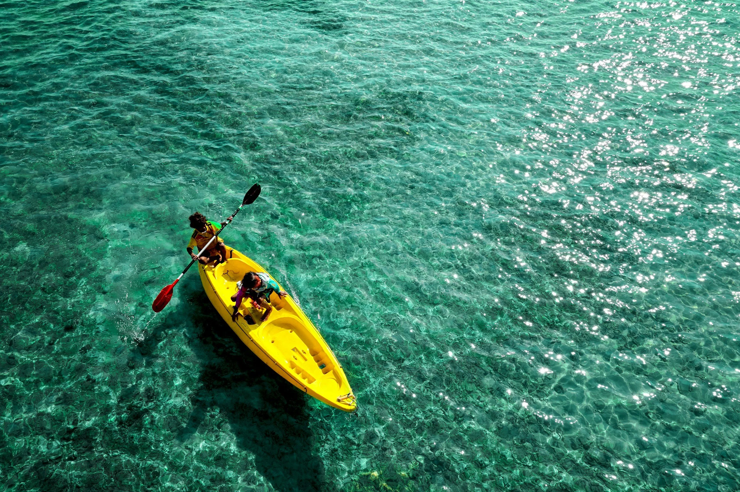 Two people kayaking in clear turquoise water, one paddling and the other sitting, in a yellow kayak.