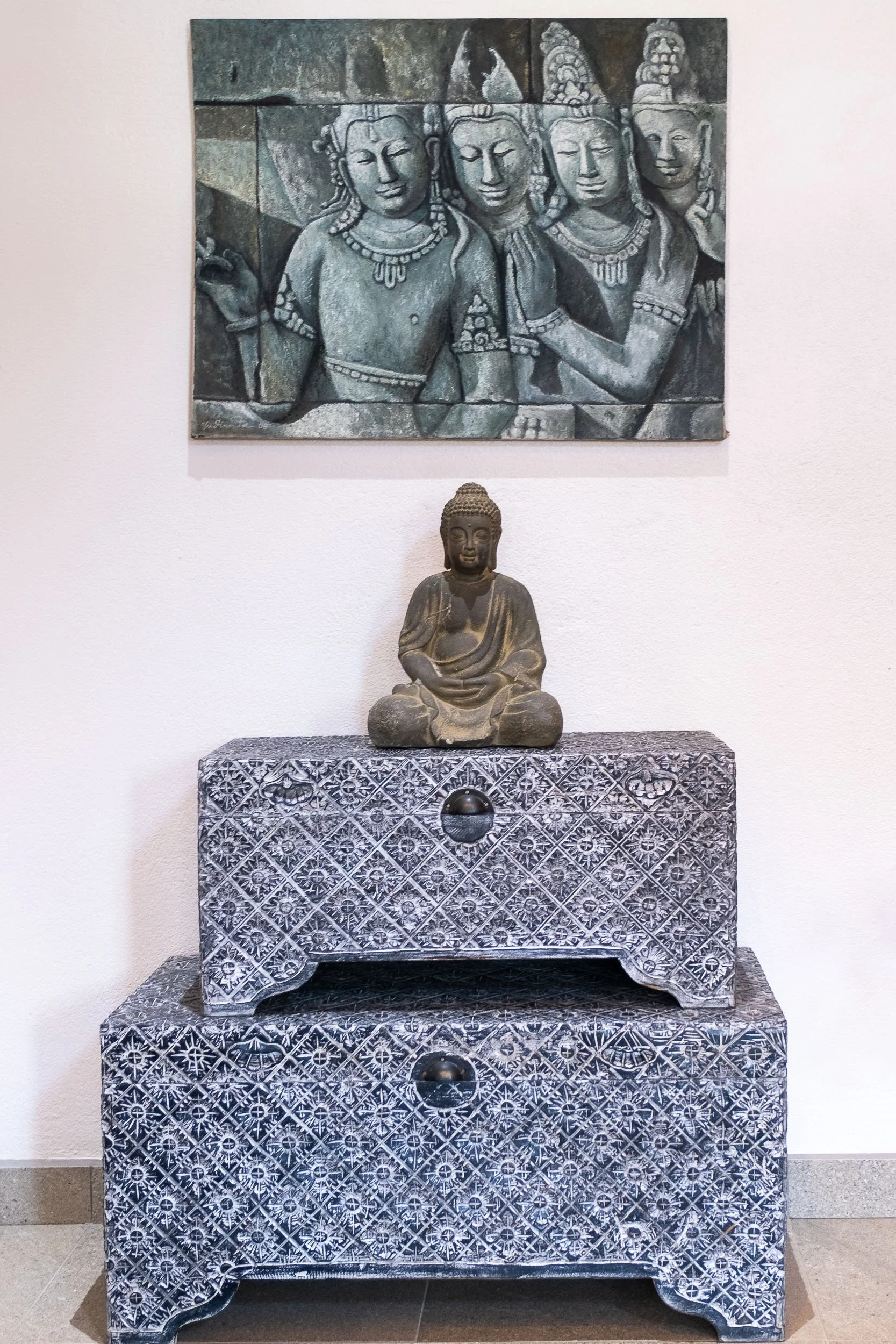 Buddha statue sitting on a patterned table, with a carved stone relief of four Hindu deities hanging on the wall above.