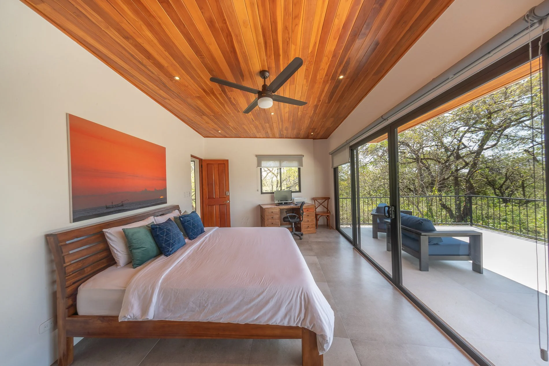 A bedroom with a wooden ceiling, ceiling fan, large sliding glass door, and balcony with outdoor seating