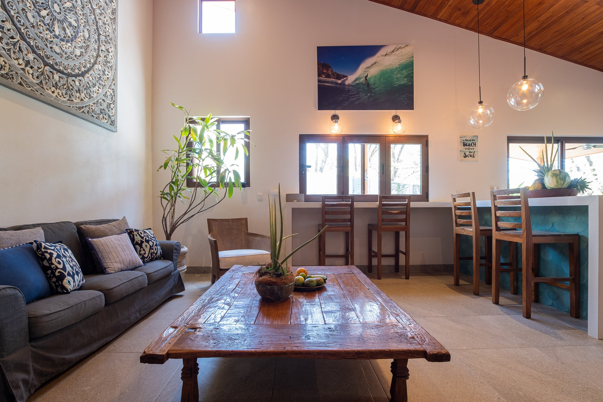 Interior of a living room with a gray couch, decorative pillows, a wooden coffee table with a potted plant, a bar area with stools, and wall art including a large surfing picture and a smaller beach-themed sign.