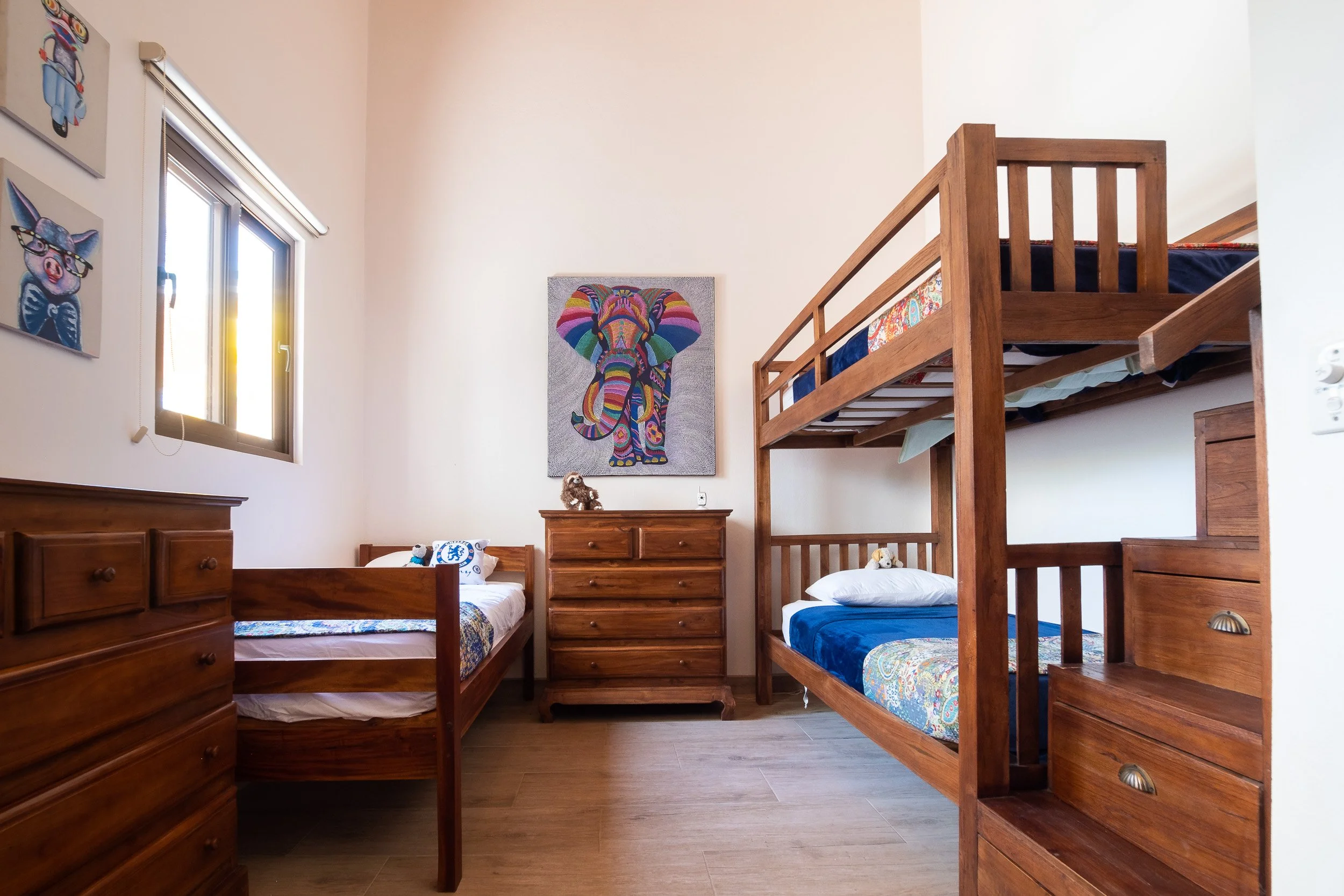 Kids' bedroom with wooden bunk beds, a single bed, wooden dressers, colorful artwork of animals on the wall, and a window letting in natural light.